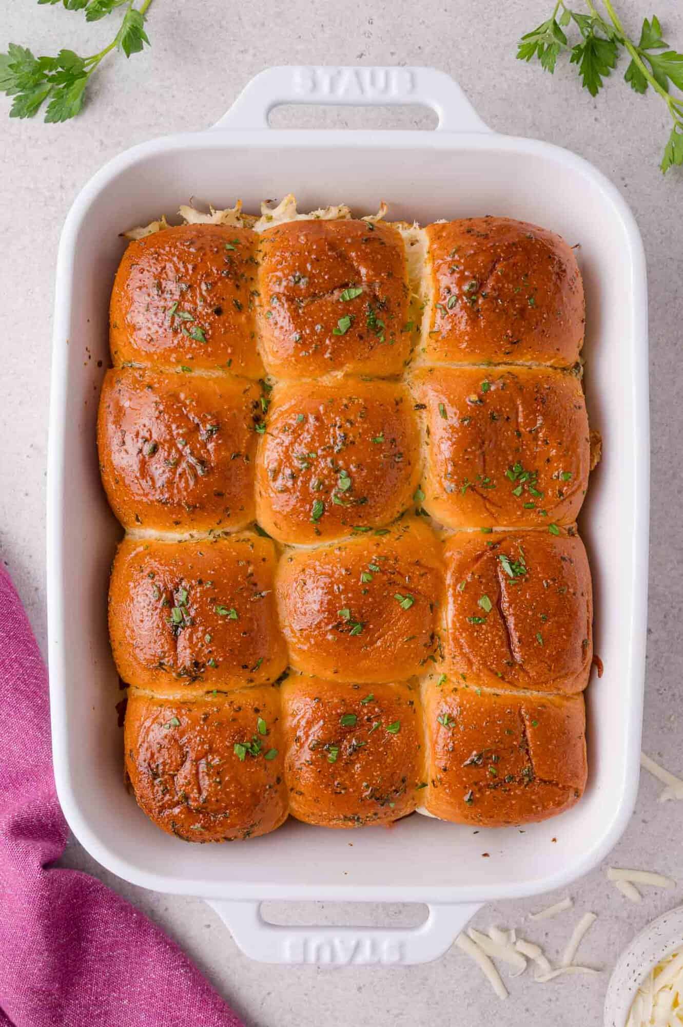 A white baking dish contains twelve golden-brown dinner rolls topped with herbs, set on a light countertop next to a purple cloth and some parsley.