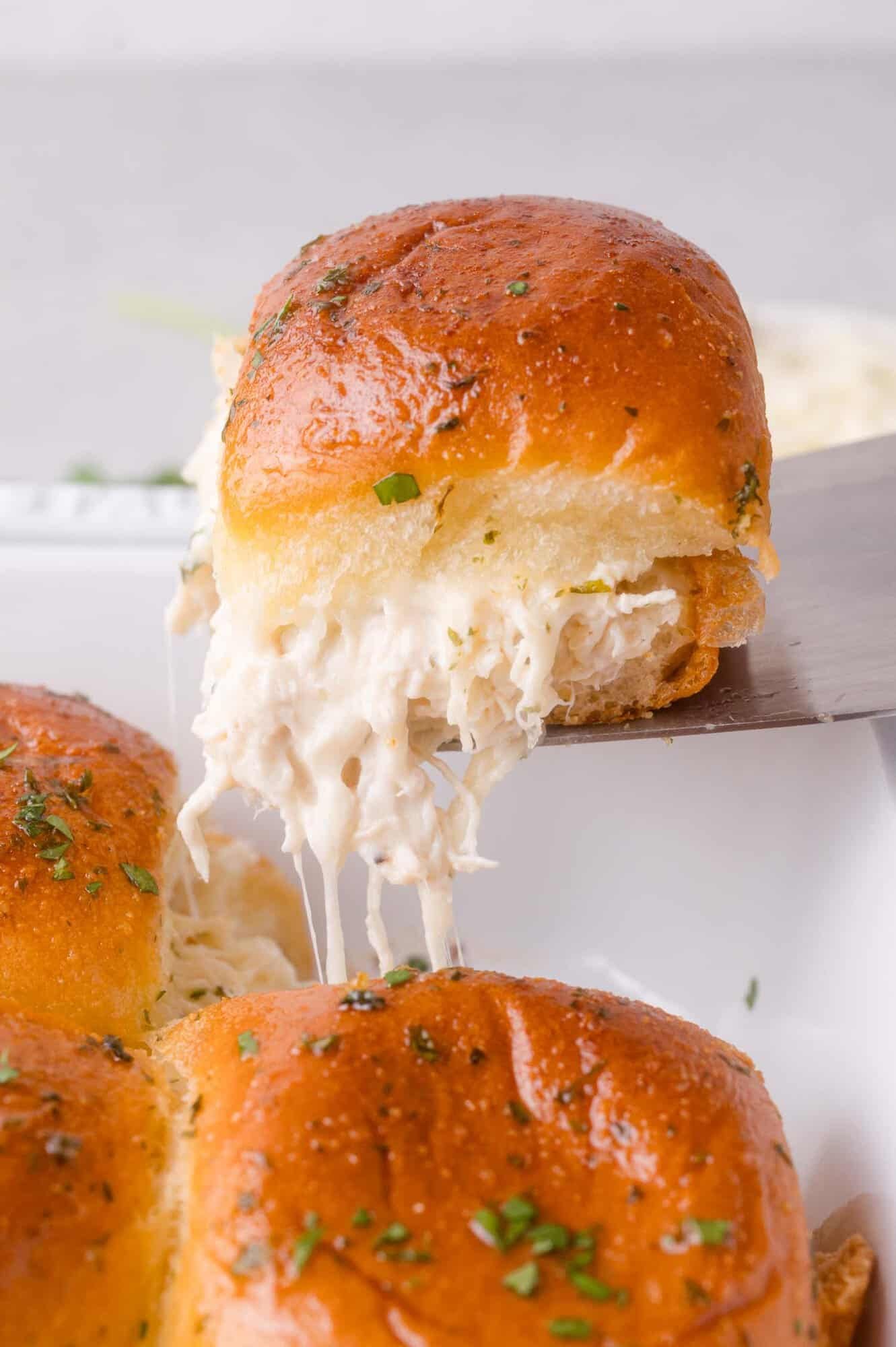 A close-up of a cheesy chicken ranch slider being lifted from a tray, with melted cheese stretching between the buns and chopped herbs sprinkled on top.