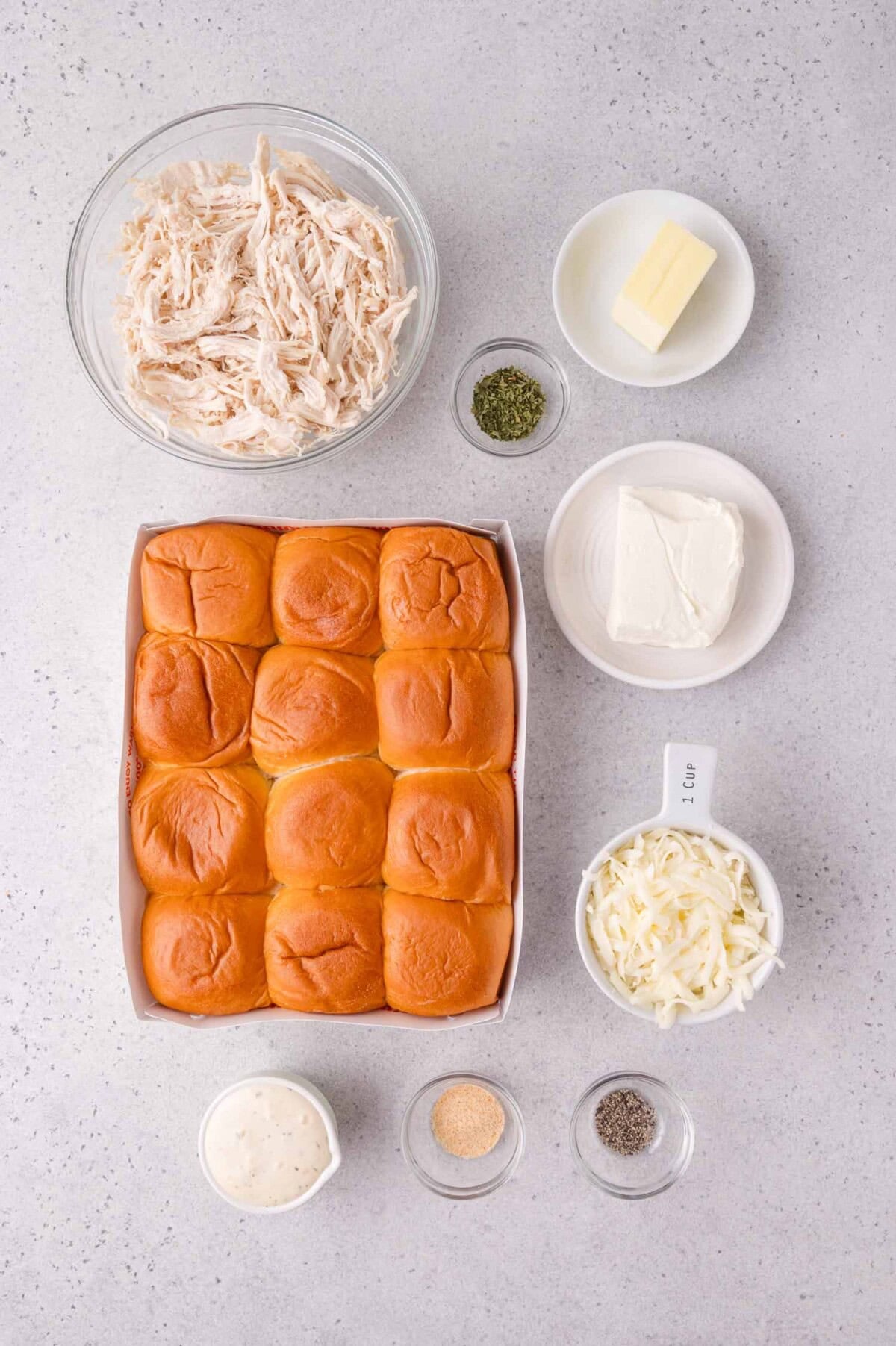 Ingredients for a chicken slider recipe, including shredded chicken, butter, cream cheese, slider buns, shredded cheese, ranch dressing, and seasonings on a light countertop.