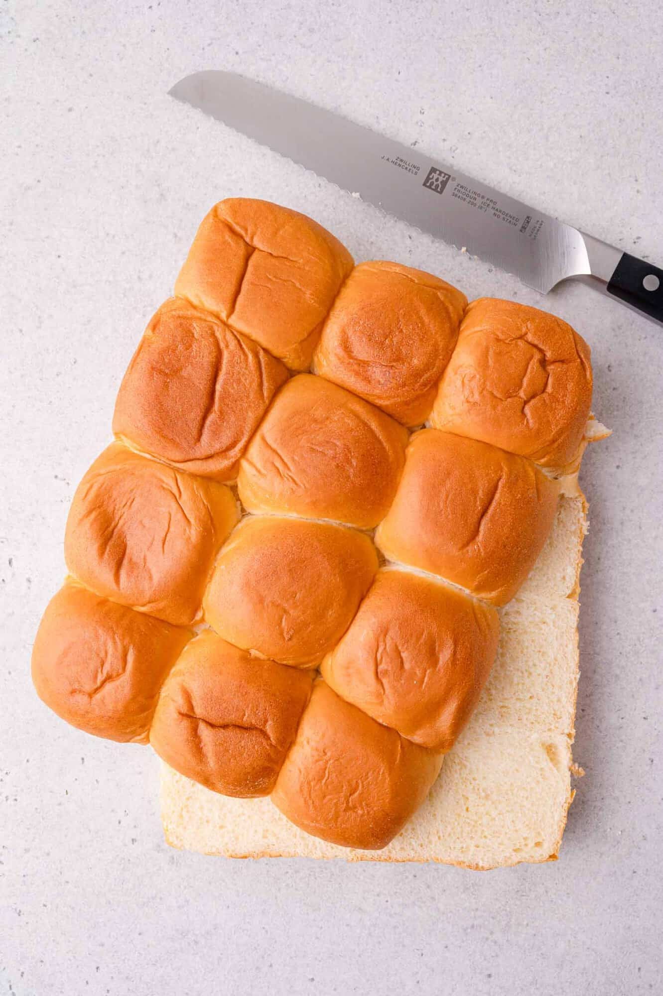 A loaf of pull-apart bread rolls, with one layer cut off at the bottom, sits on a light surface next to a bread knife.