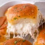 A golden-brown slider bun filled with melted, stretchy cheese and chicken is being lifted from a pan, garnished with chopped herbs.