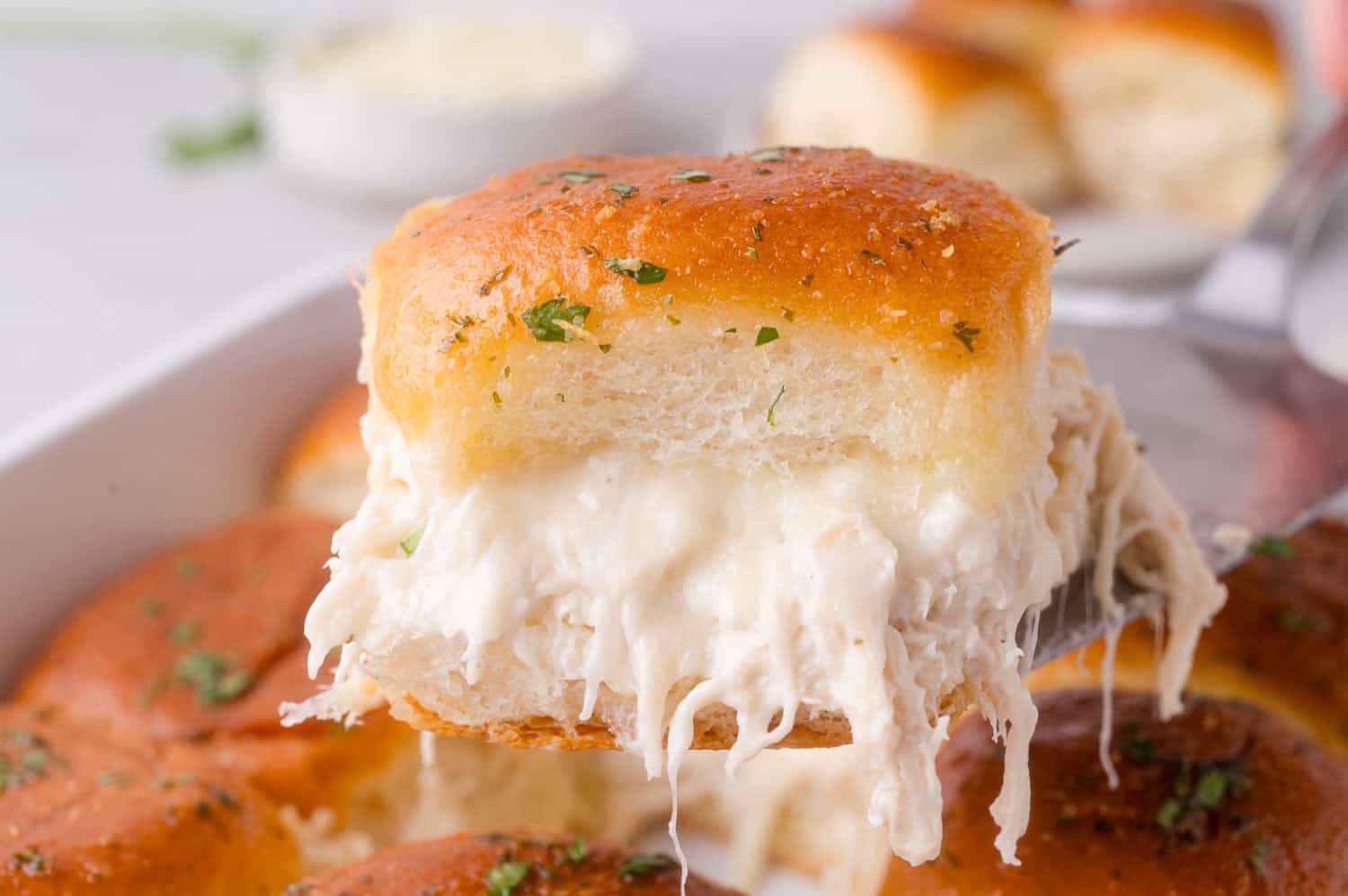 A close-up of a cheesy chicken slider on a spatula, topped with herbs, showing creamy shredded chicken filling and golden brown bread.