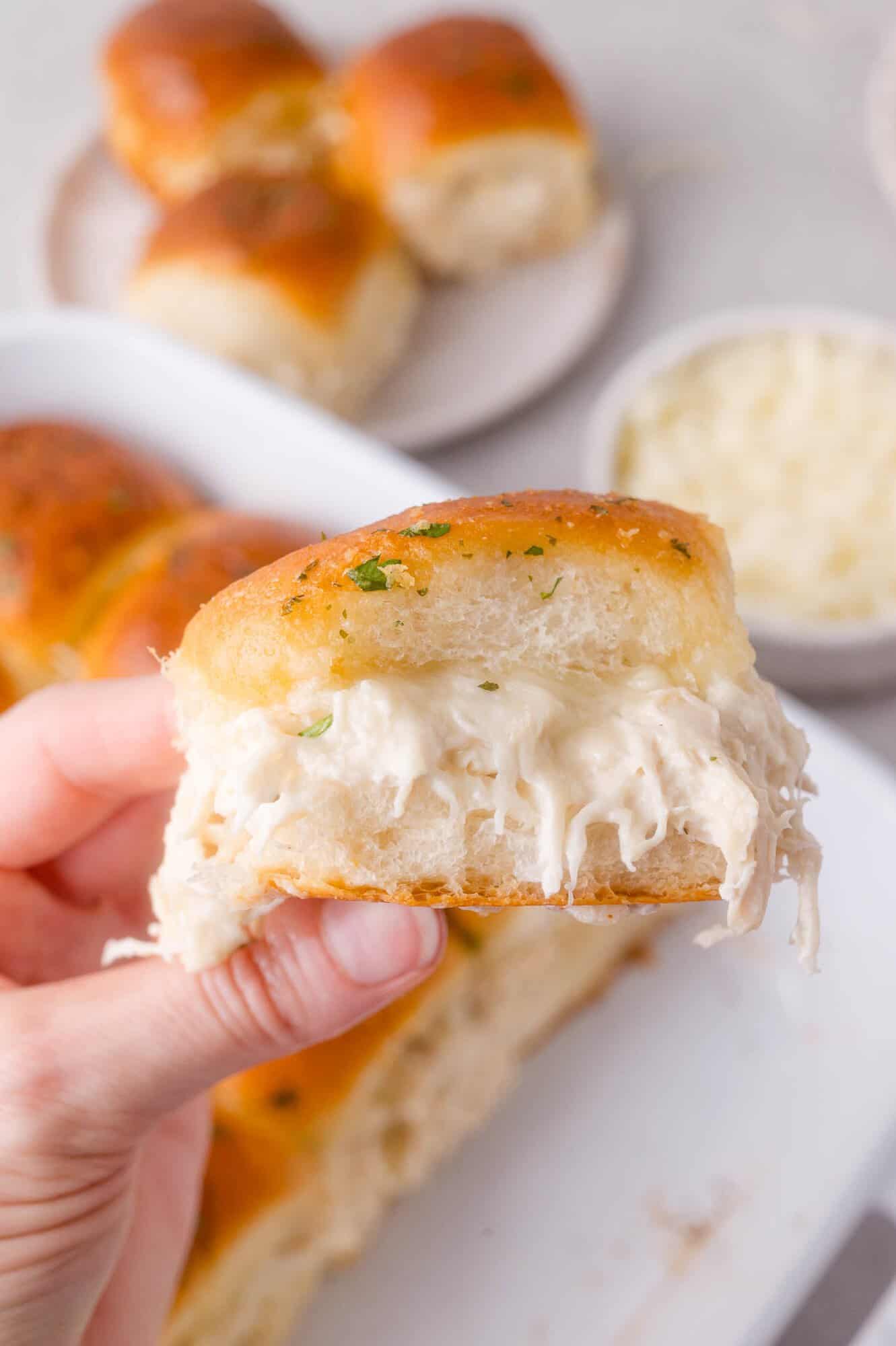 A hand holds a slider filled with creamy shredded ranch chicken between two golden brown rolls; more sliders and a bowl of cheese are in the background.