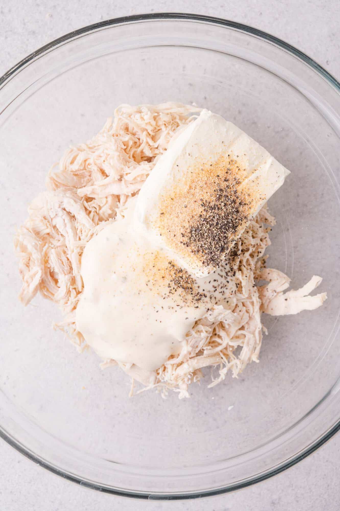 A glass bowl containing shredded chicken, cream cheese, ranch dressing, and seasonings on a light countertop.