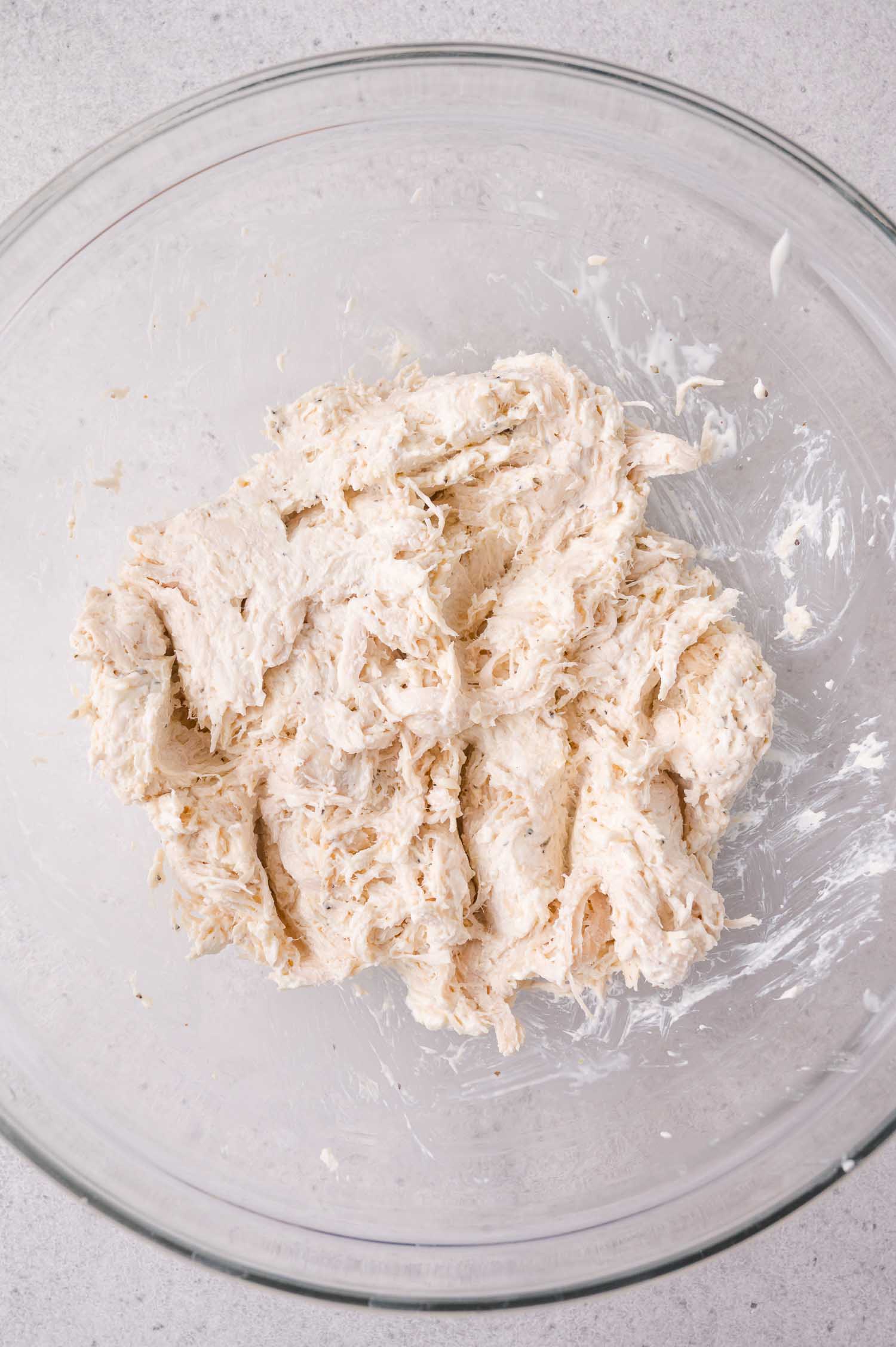 Shredded chicken in a clear glass mixing bowl on a light-colored surface.