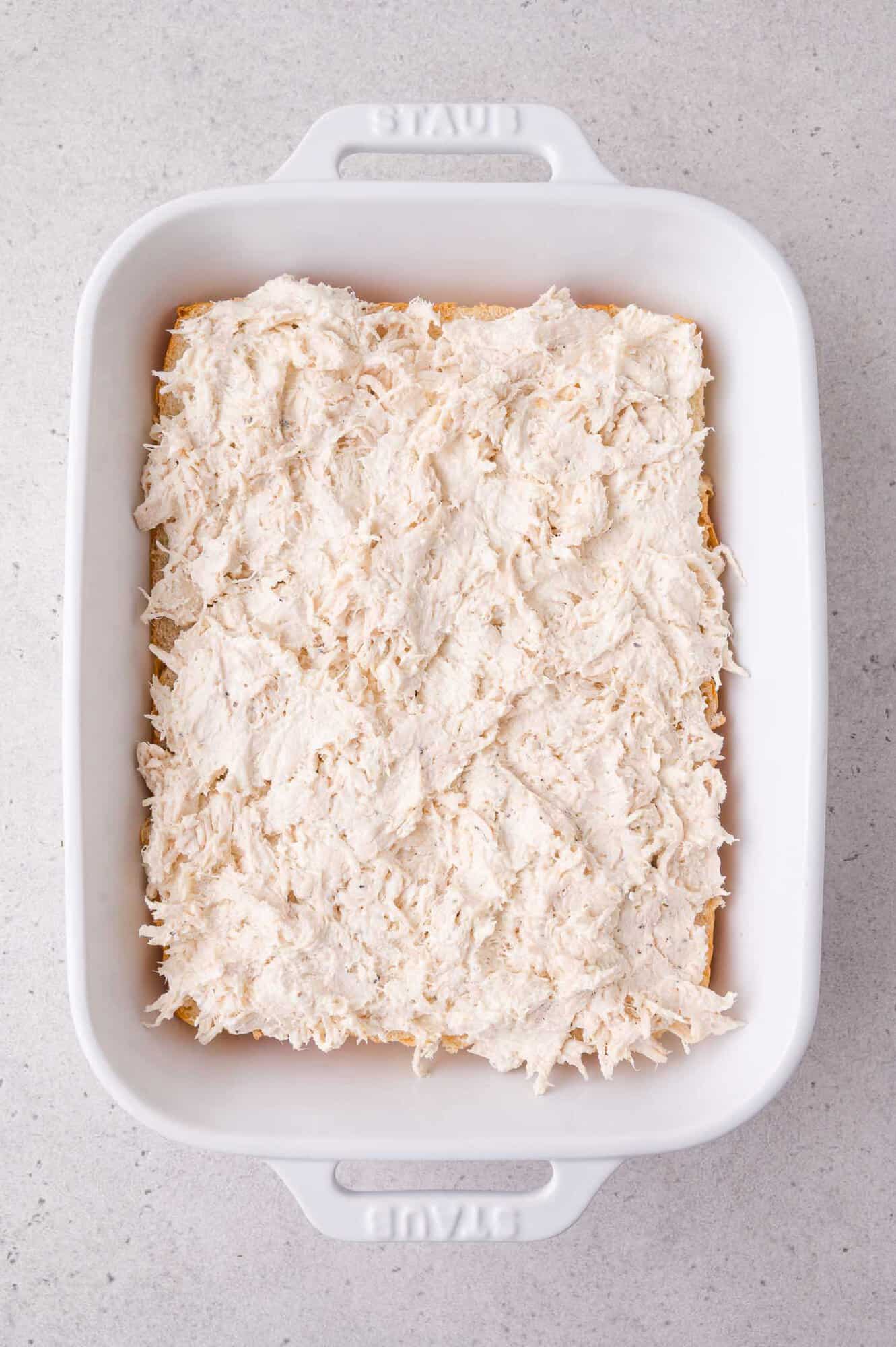 A white baking dish filled with a layer of shredded chicken spread evenly over a base of slider buns, on a light-colored countertop.