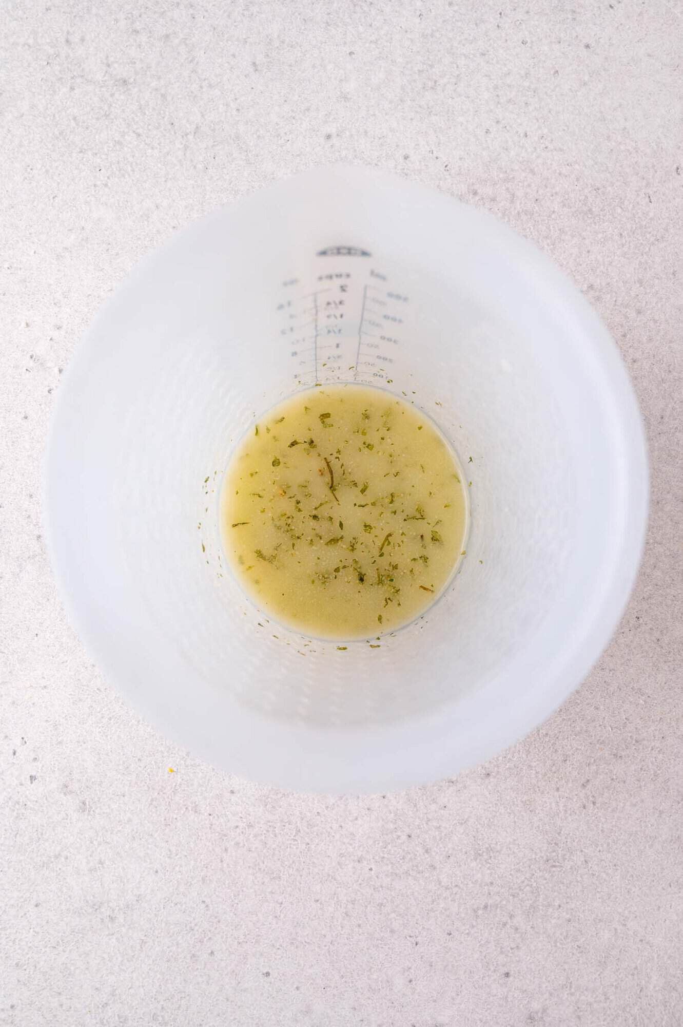 A small amount of melted butter with herbs sits at the bottom of a clear plastic measuring cup on a light-colored surface.