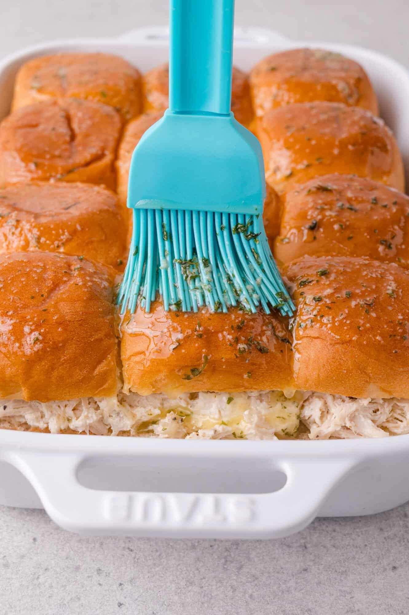 A blue brush spreads melted herb butter on baked slider rolls filled with shredded chicken in a white baking dish.