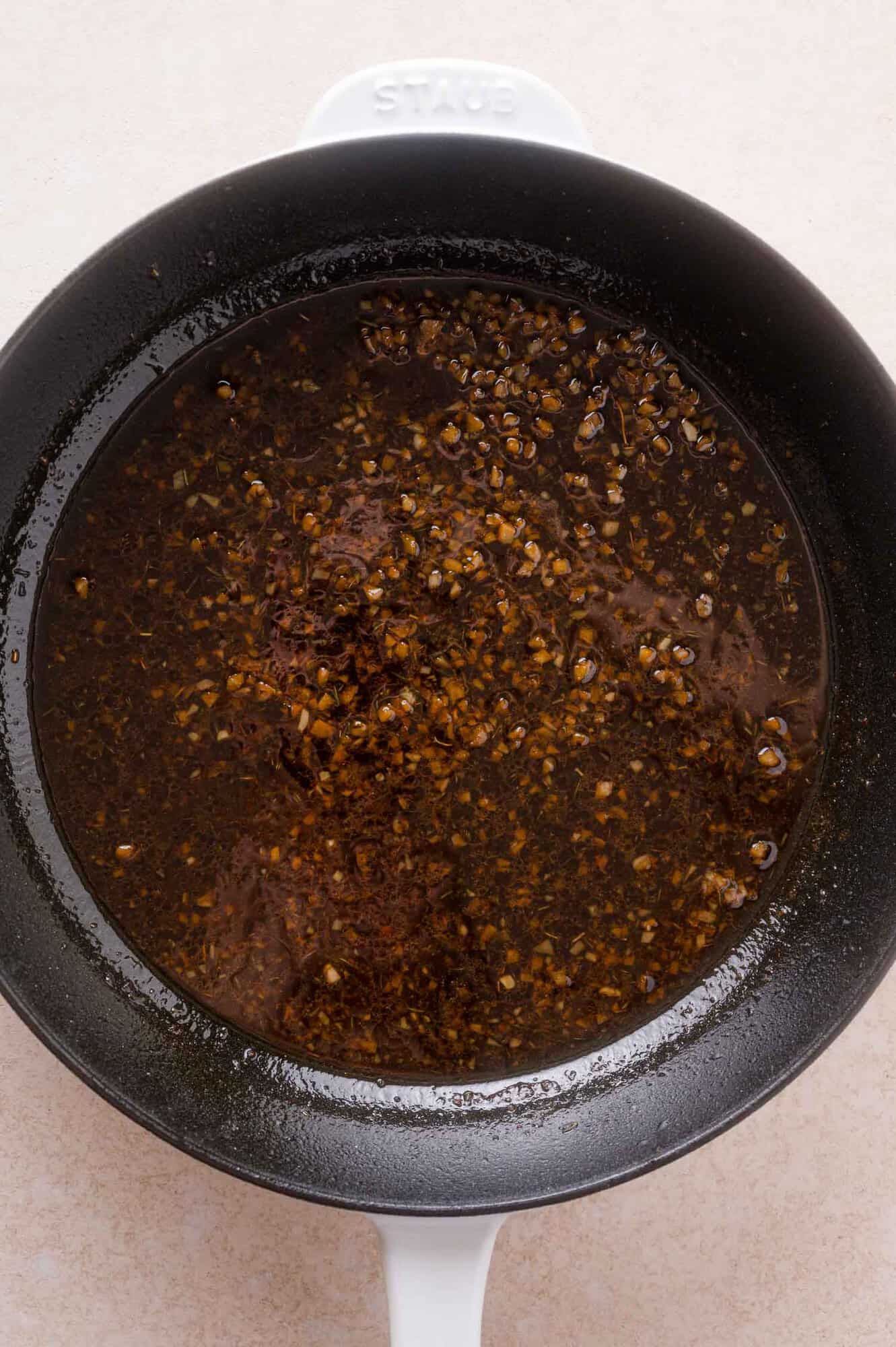 A black pan with finely chopped garlic and dark sauce simmering on the stovetop, viewed from above.