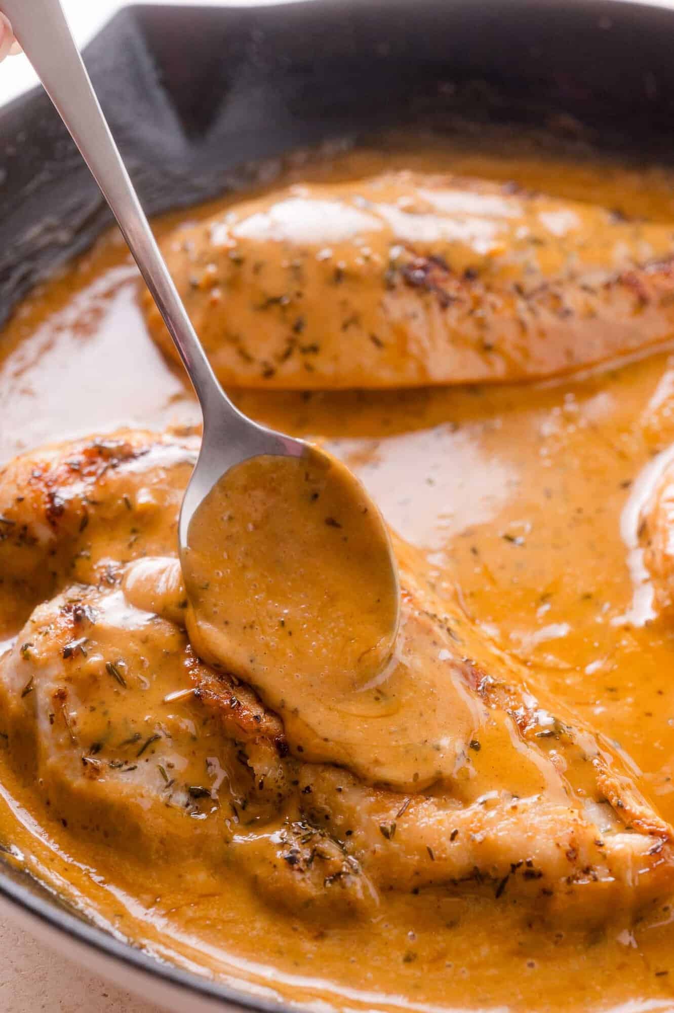 A spoon pours creamy herb sauce over seared chicken breasts in a black skillet.