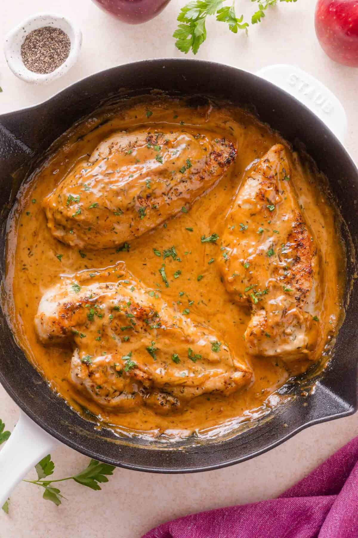 Three cooked chicken breasts in a cast iron skillet with apple cider cream sauce, garnished with chopped herbs.