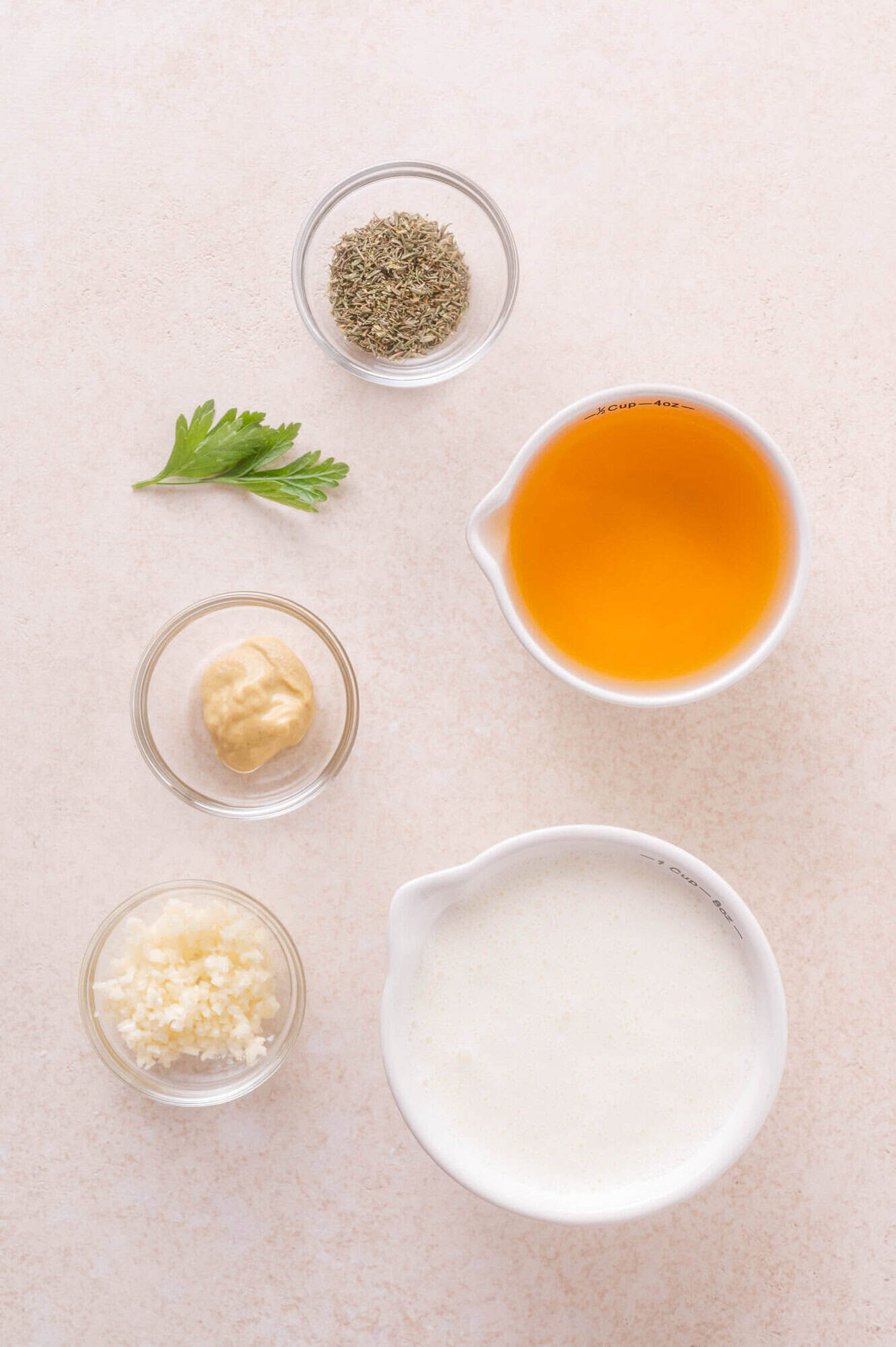 Five ingredients are arranged on a light surface: dried herbs, apple cider, Dijon mustard, minced garlic, cream, with a parsley leaf for garnish.