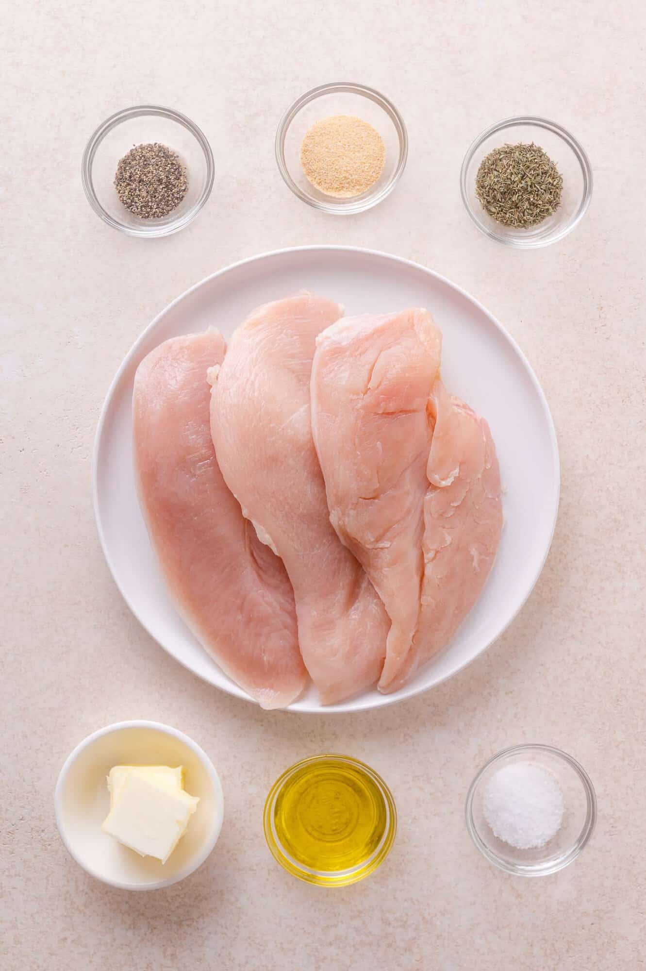 Raw chicken breasts on a white plate surrounded by small bowls of black pepper, garlic powder, dried herbs, butter, olive oil, and salt on a light surface.