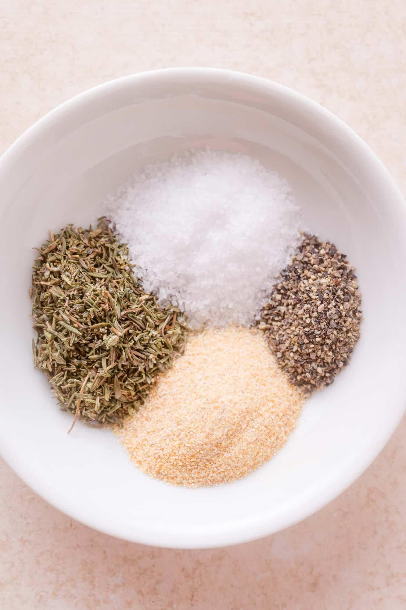 A white bowl containing four spices: dried herbs, coarse salt, ground black pepper, and garlic powder, arranged in separate sections.