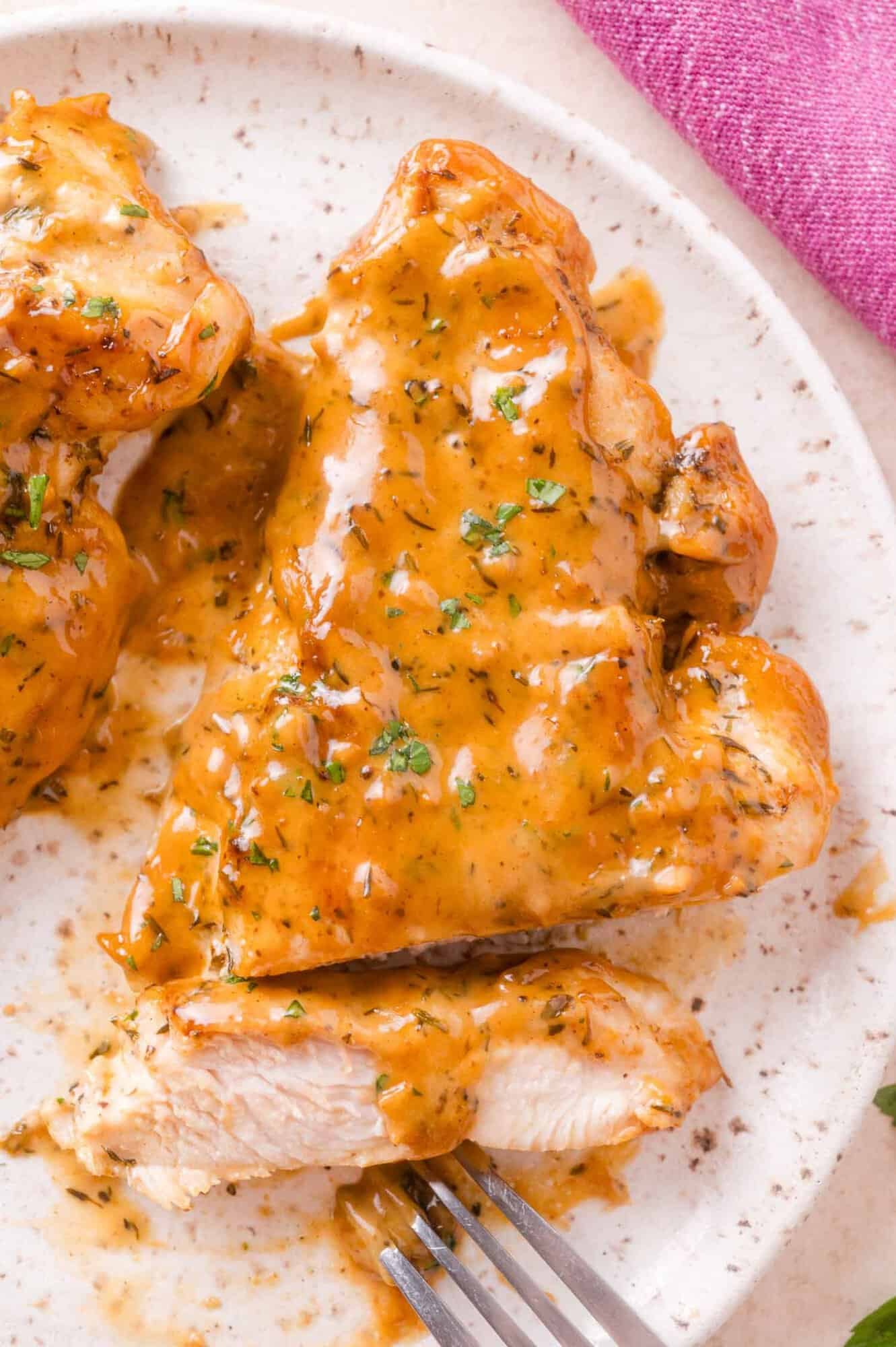 A cooked chicken breast on a plate covered in apple cider cream sauce, with a fork and a piece cut off; a pink cloth is partly visible.