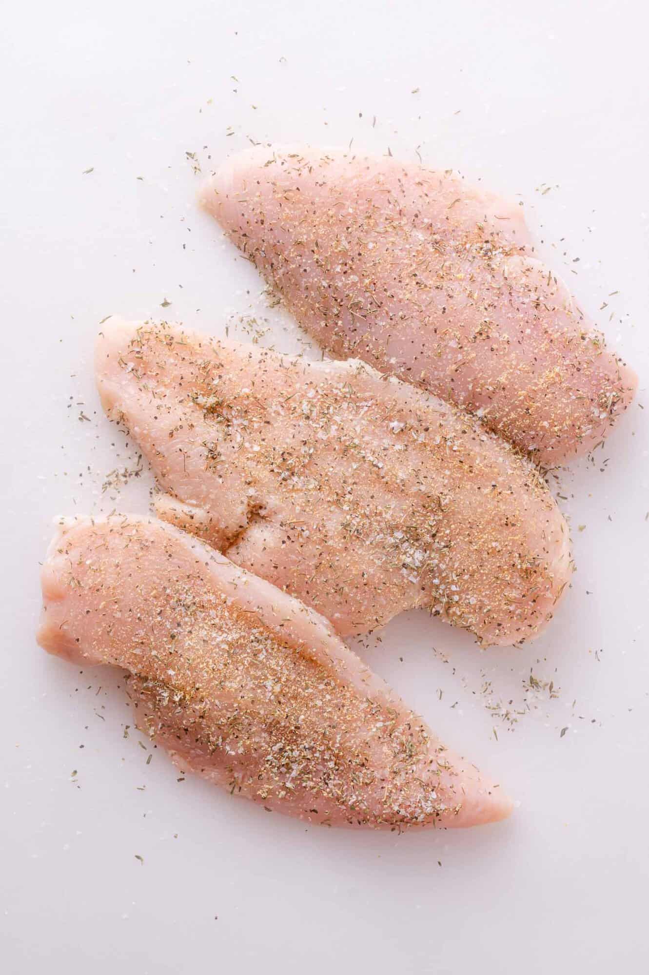Three raw chicken breasts on a white surface, sprinkled with a mixture of dried herbs and seasonings.