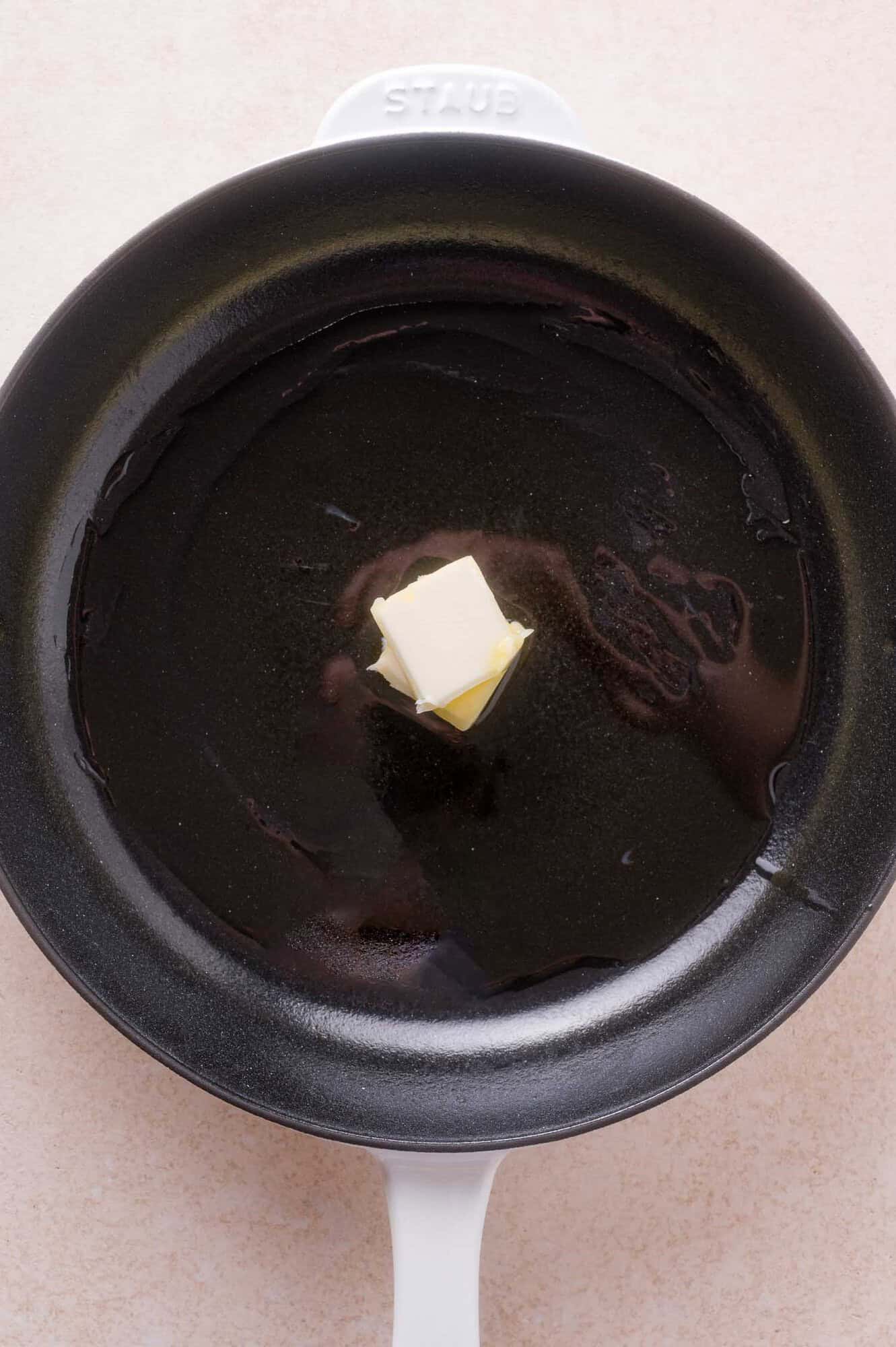 A pat of butter melting in the center of a black skillet with a white handle on a light countertop.