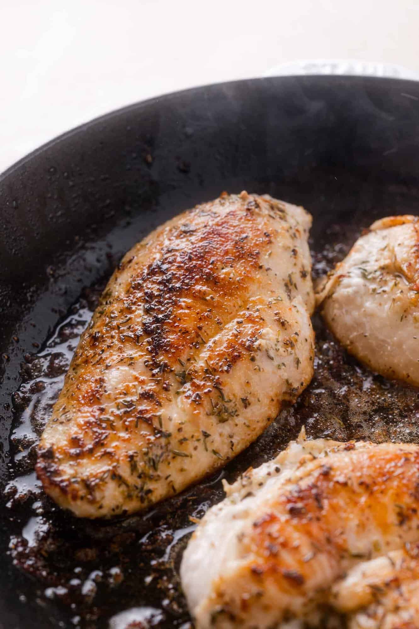 Three seasoned chicken breasts cooking in a black skillet, with browned, crispy edges and visible herbs on the surface.