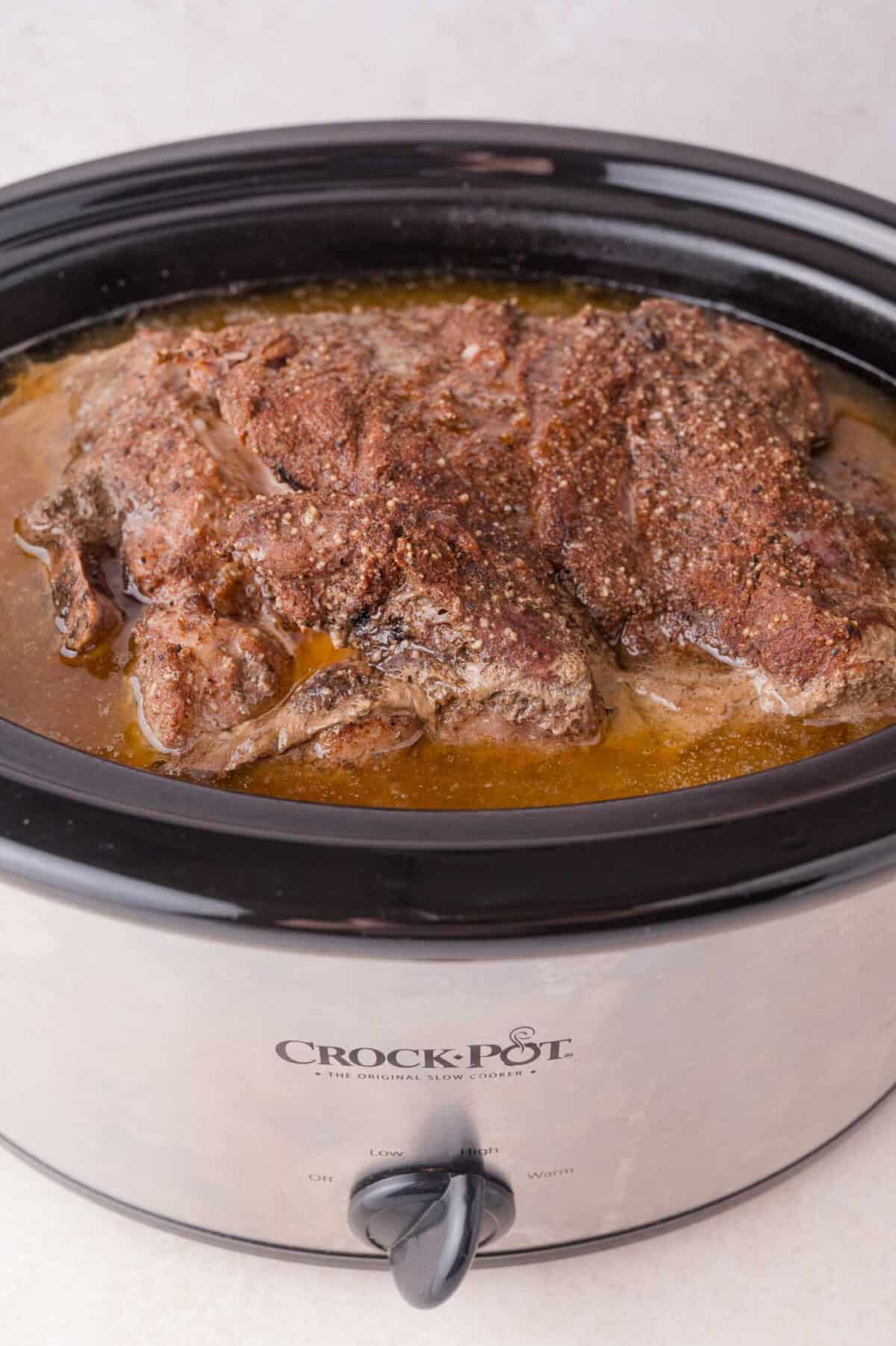 Cooked seasoned pork sits in broth inside a black Crock-Pot slow cooker on a light countertop.