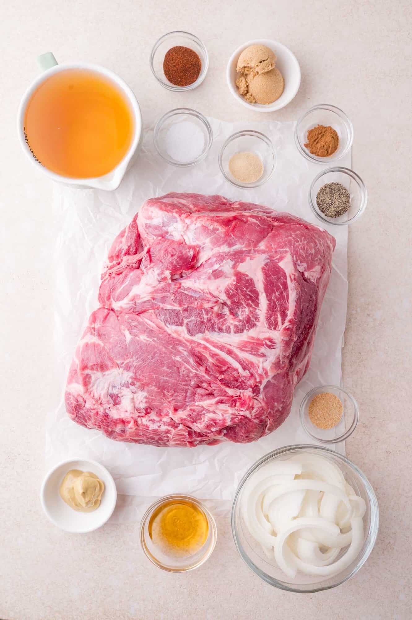 A raw pork shoulder on parchment paper is surrounded by small bowls of seasonings, sliced onions, broth, mustard, and brown sugar on a light surface.