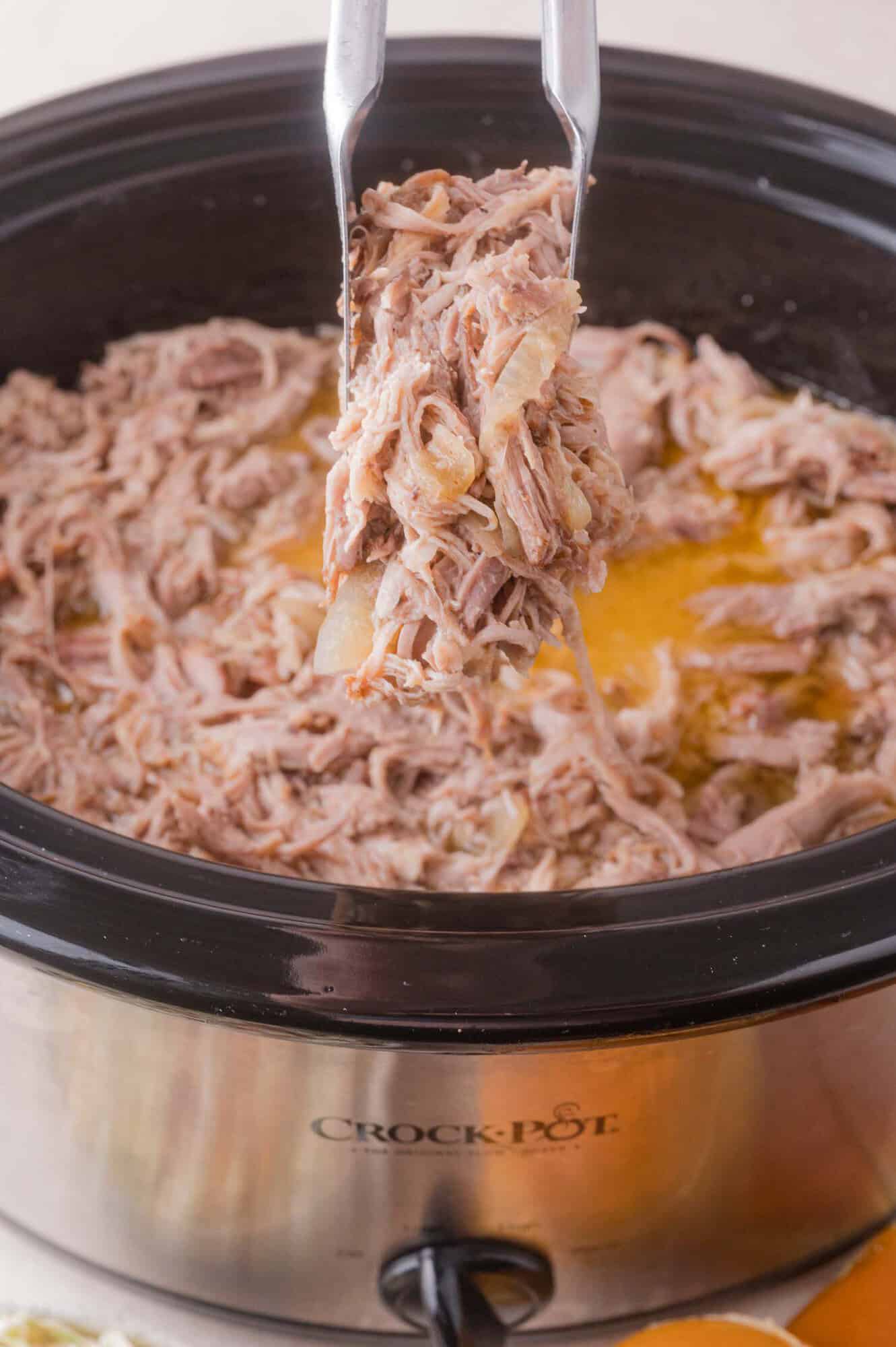 Shredded pork being lifted with tongs from a slow cooker filled with cooked meat and juices.
