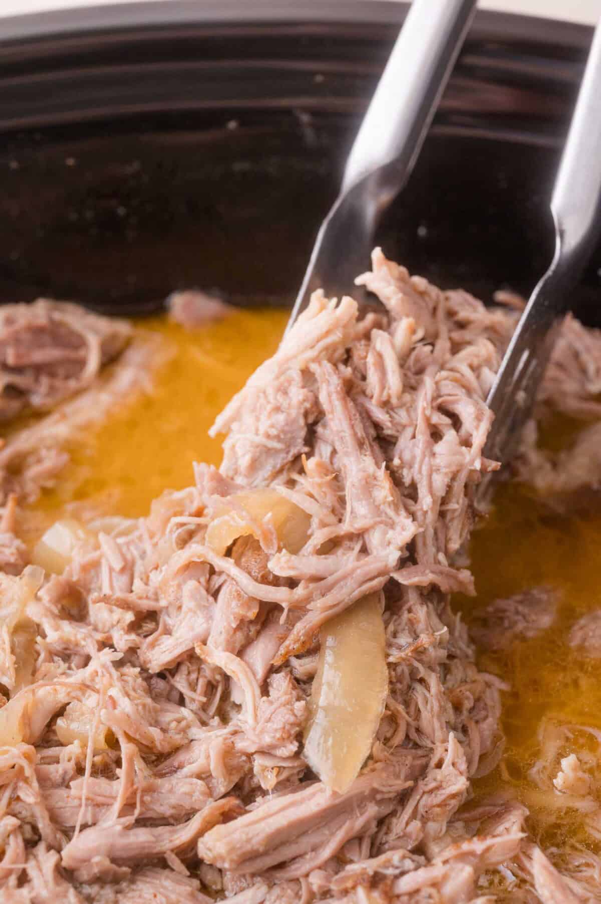 Cooked shredded pork being lifted from a slow cooker with metal tongs, surrounded by broth and pieces of onion.