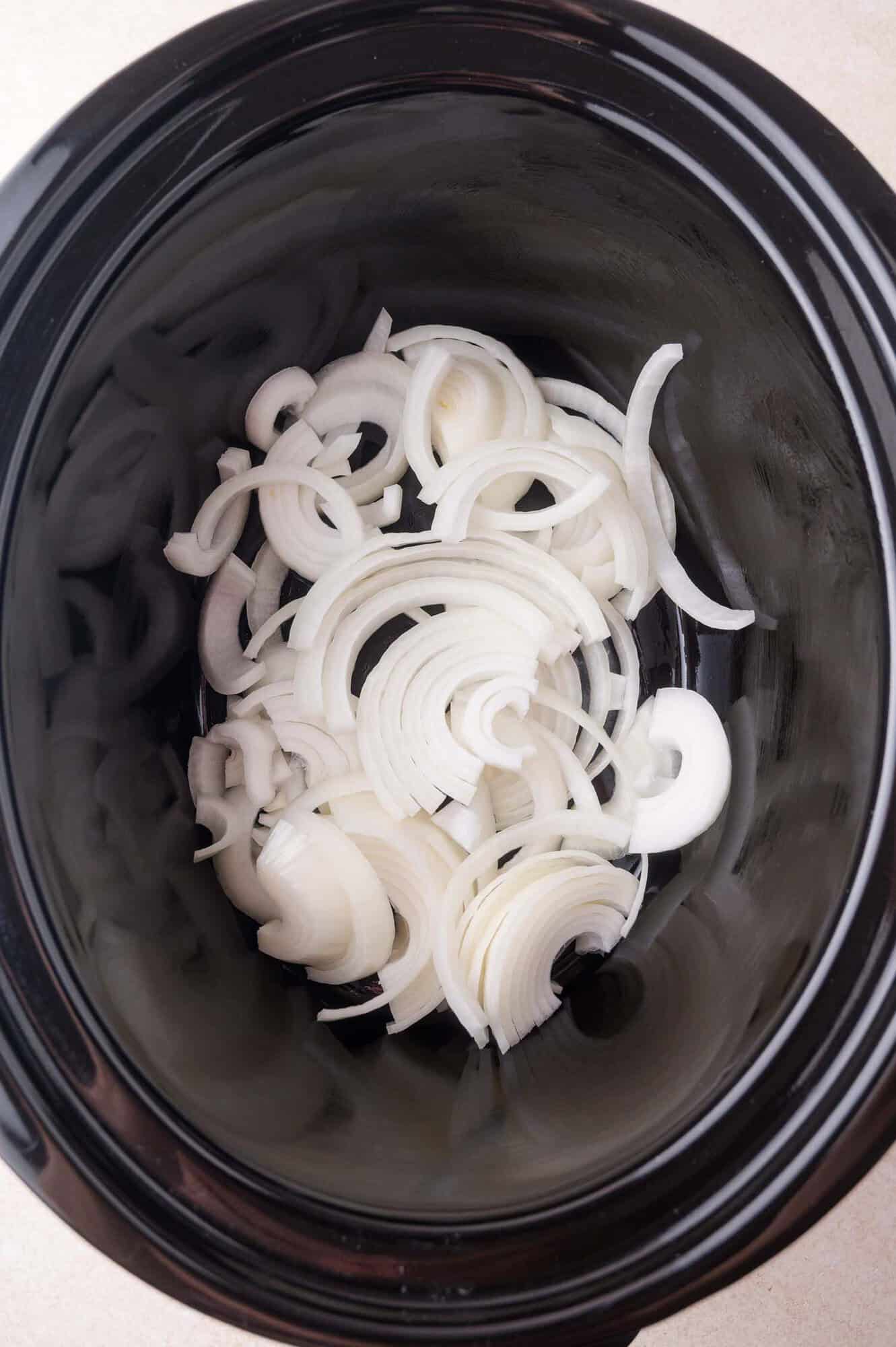 Sliced white onions spread at the bottom of a black slow cooker.