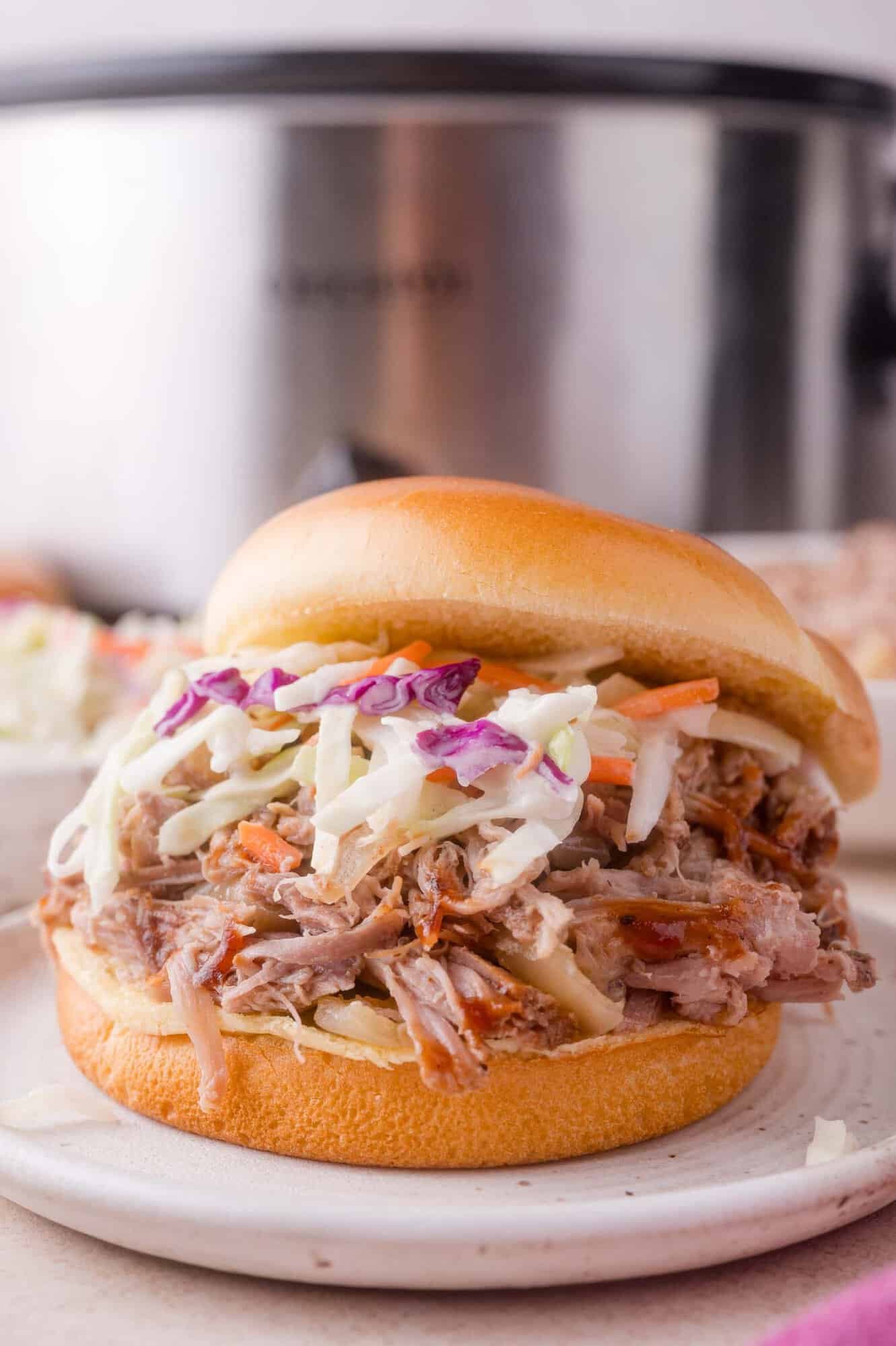 Pulled pork sandwich topped with coleslaw on a white bun, served on a plate with a slow cooker in the background.