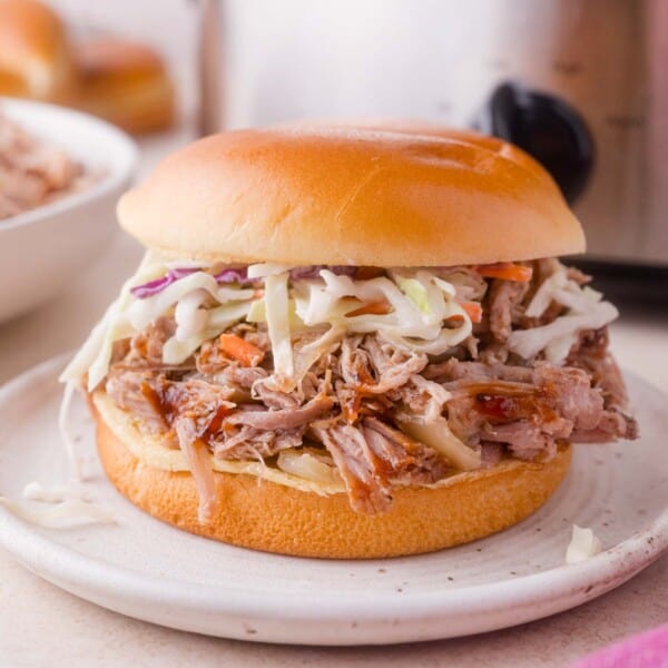 Pulled pork sandwich topped with coleslaw on a white plate, with a blurred bowl of slaw and a slow cooker in the background.