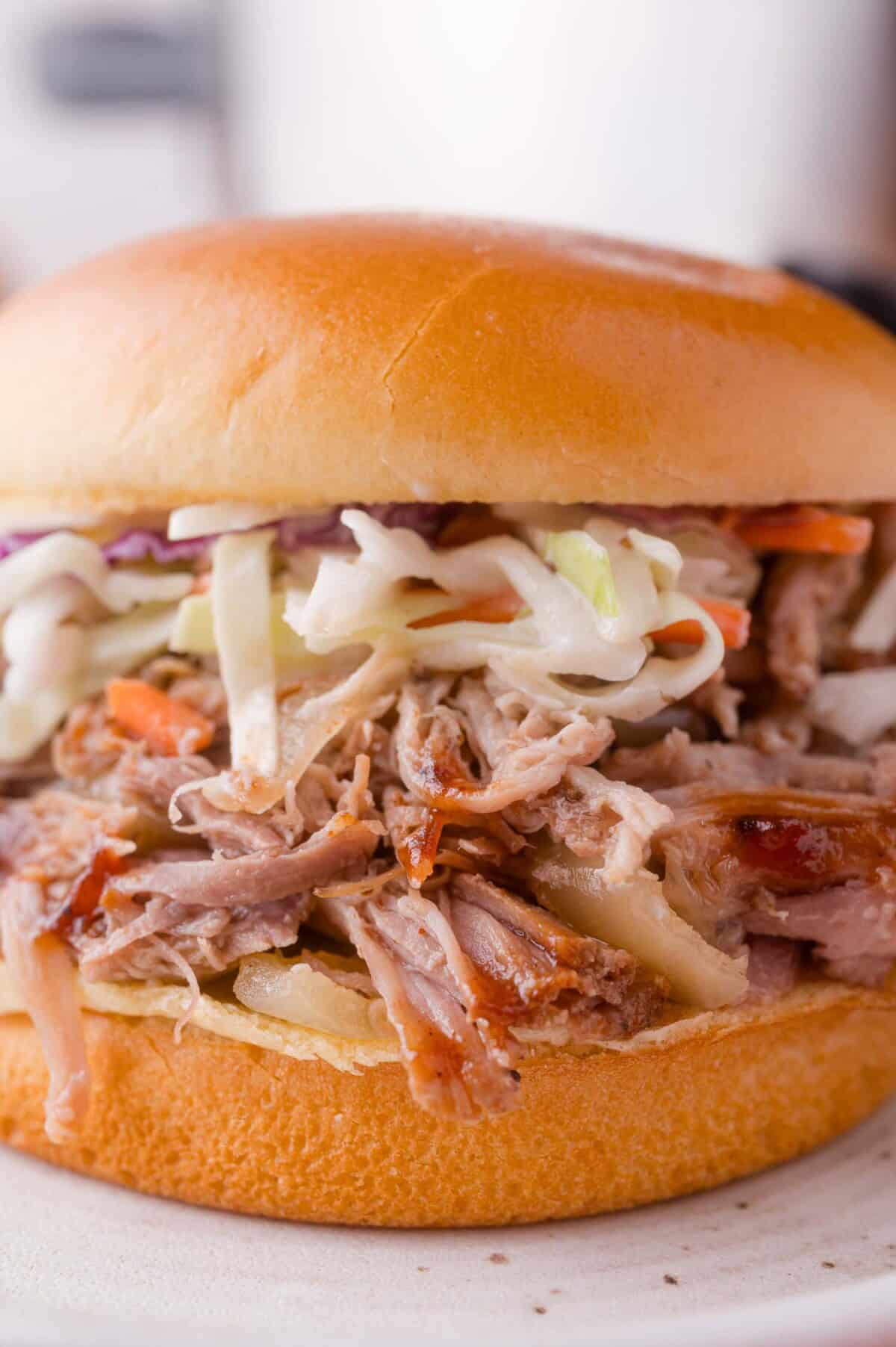Pulled pork sandwich topped with coleslaw on a soft hamburger bun, served on a white plate.