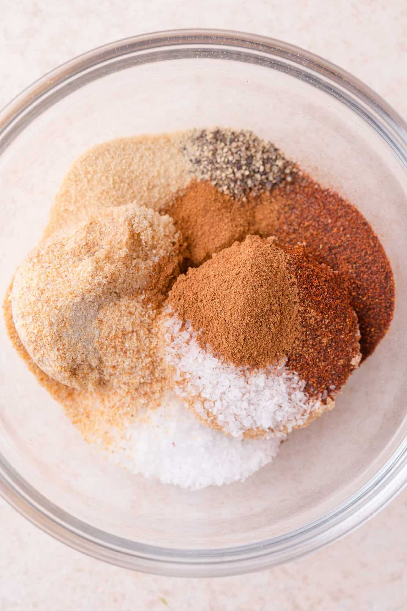 A glass bowl filled with various ground spices and coarse salt, arranged in small piles on a light surface.