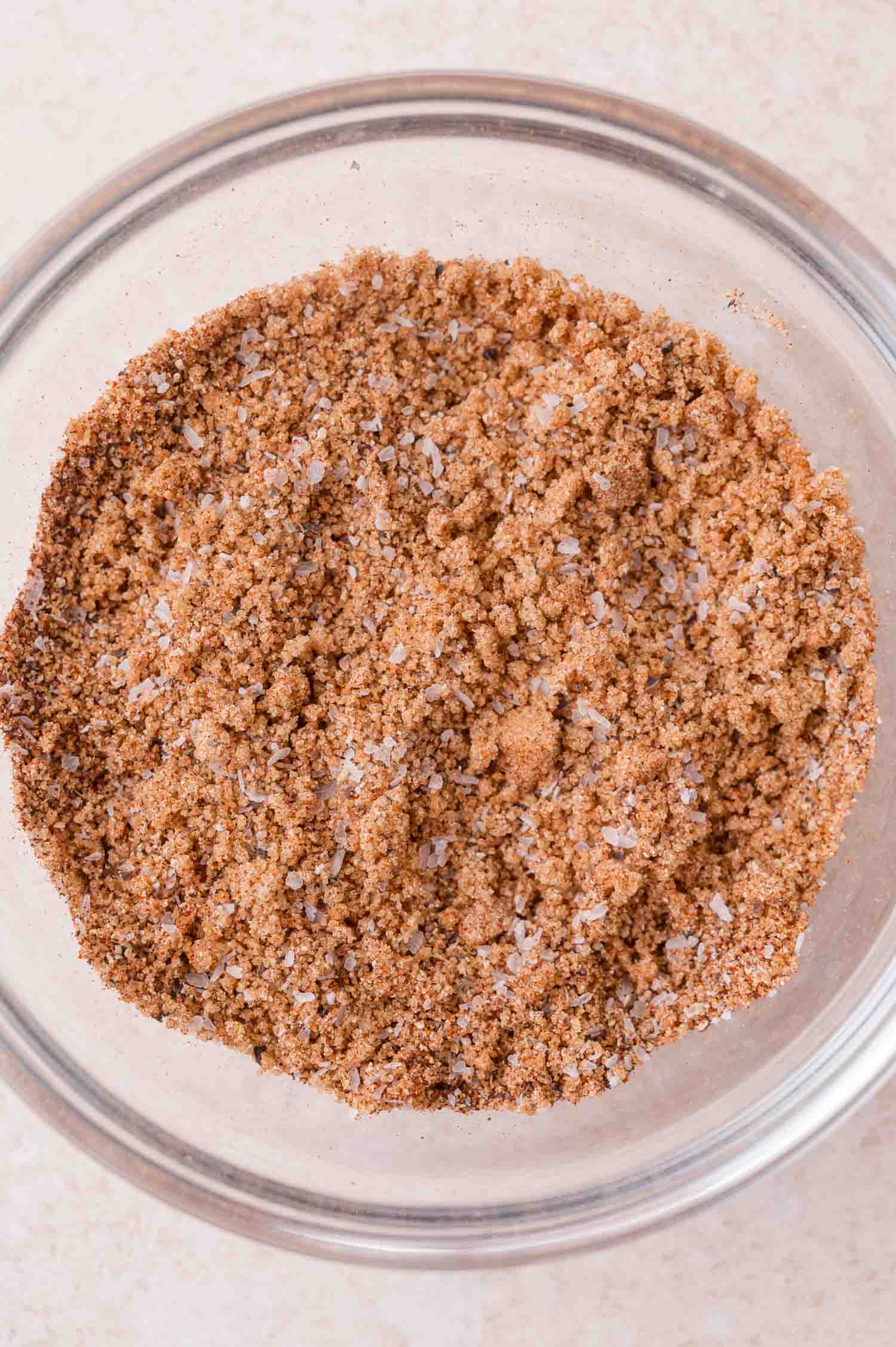A glass bowl filled with a coarse brown spice dry rub, on a light-colored surface.