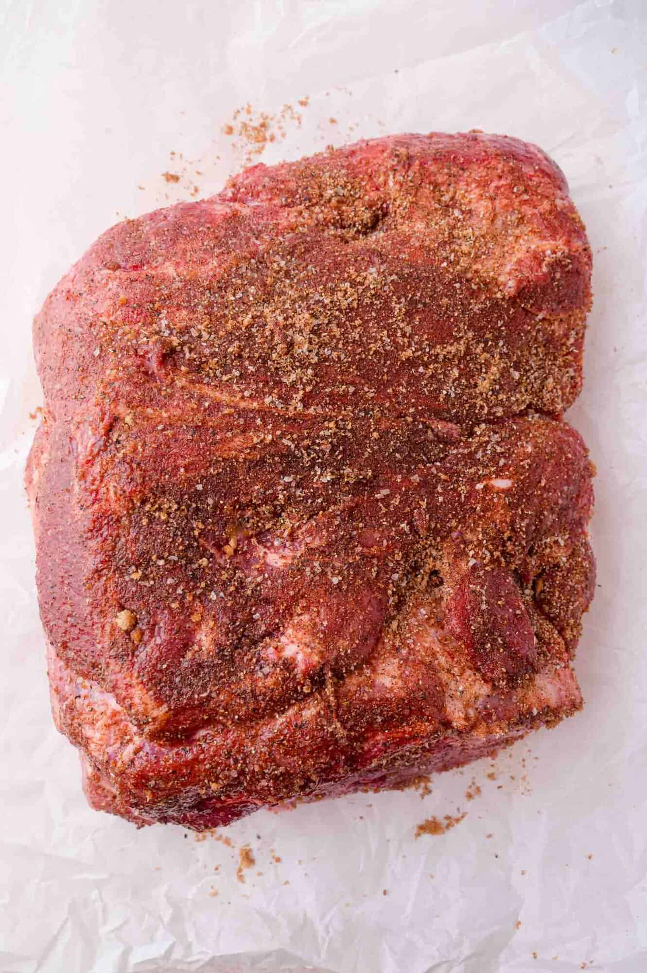 A raw cut of pork covered with a dry spice rub sits on a sheet of white parchment paper.