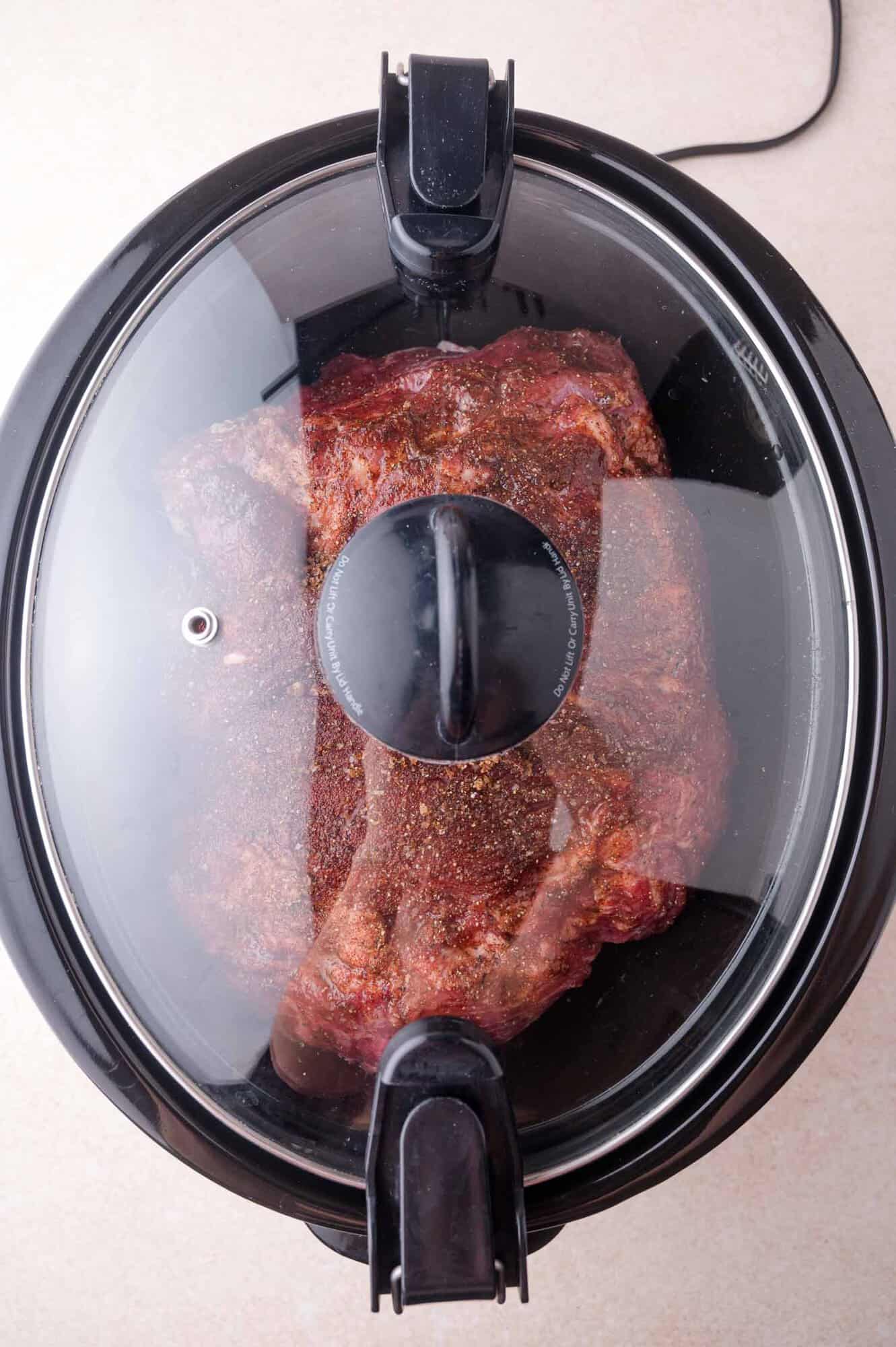 A seasoned piece of meat is visible through the glass lid of a black slow cooker on a light countertop.