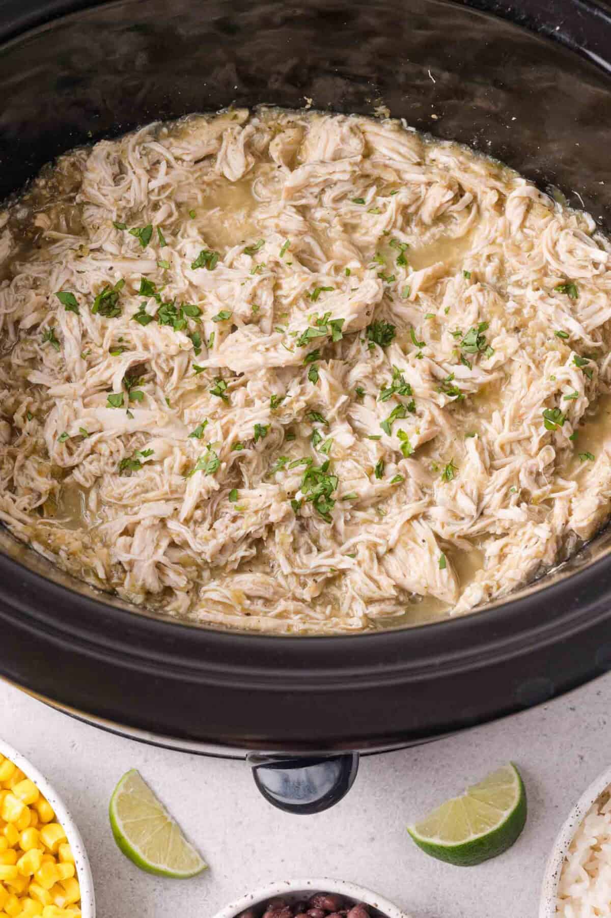 Shredded chicken with salsa verde in a slow cooker, surrounded by small bowls of corn, black beans, and lime wedges.