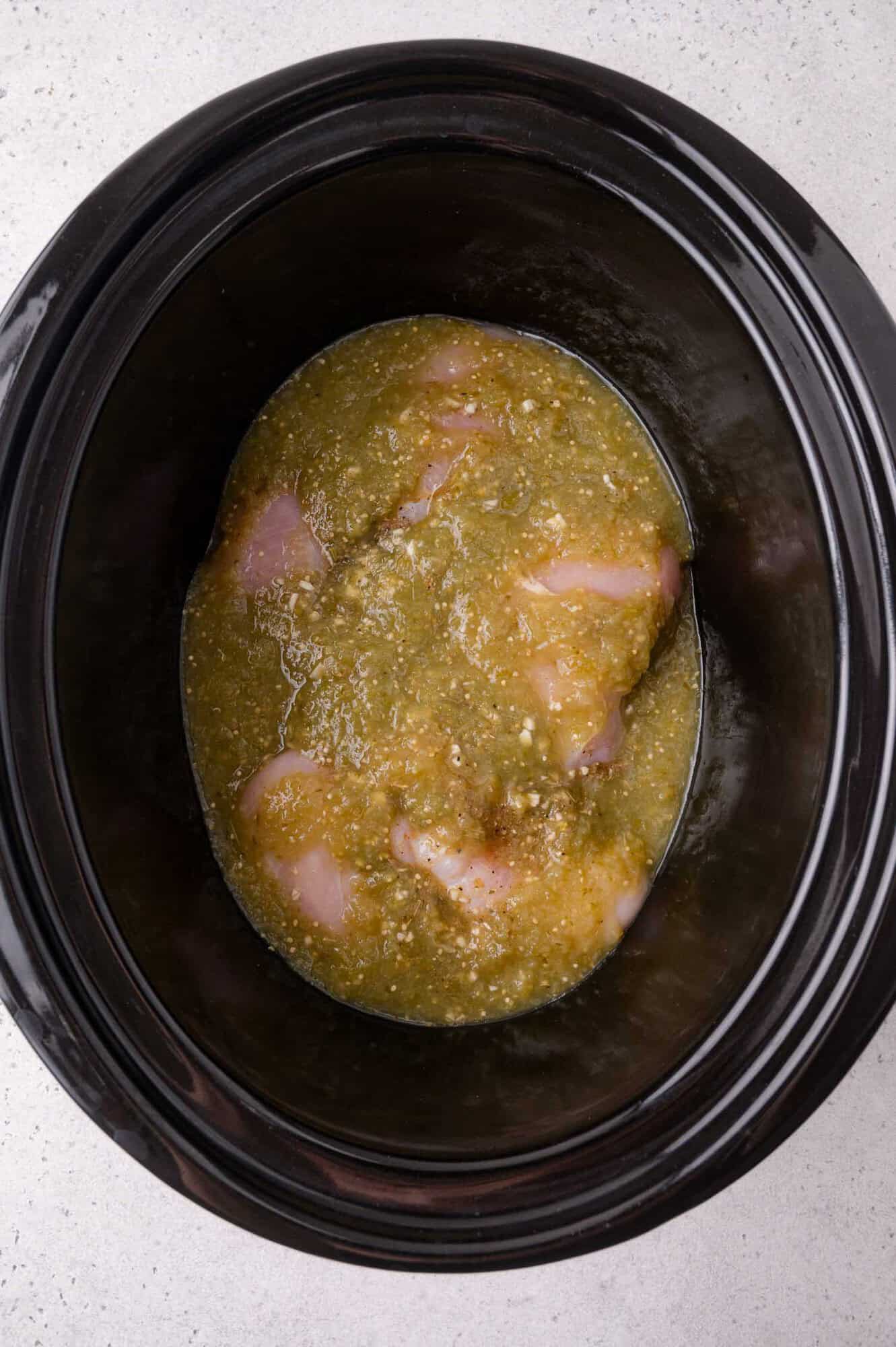 Raw chicken breasts covered in salsa verde inside a black slow cooker, photographed from above on a light-colored surface.