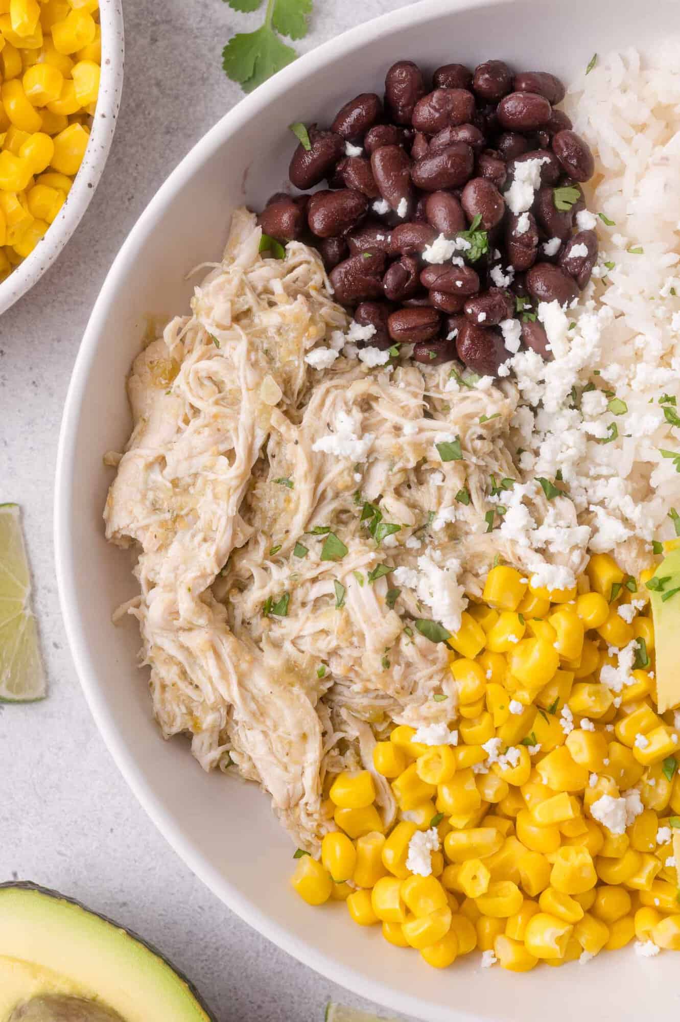 A bowl with shredded slow cooker salsa verde chicken, black beans, corn, white rice, avocado, and crumbled cheese, garnished with herbs.