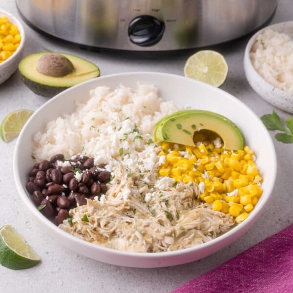 A bowl containing slow cooker salsa verde chicken, white rice, black beans, corn, avocado slices, and crumbled cheese, with lime wedges and a slow cooker in the background.