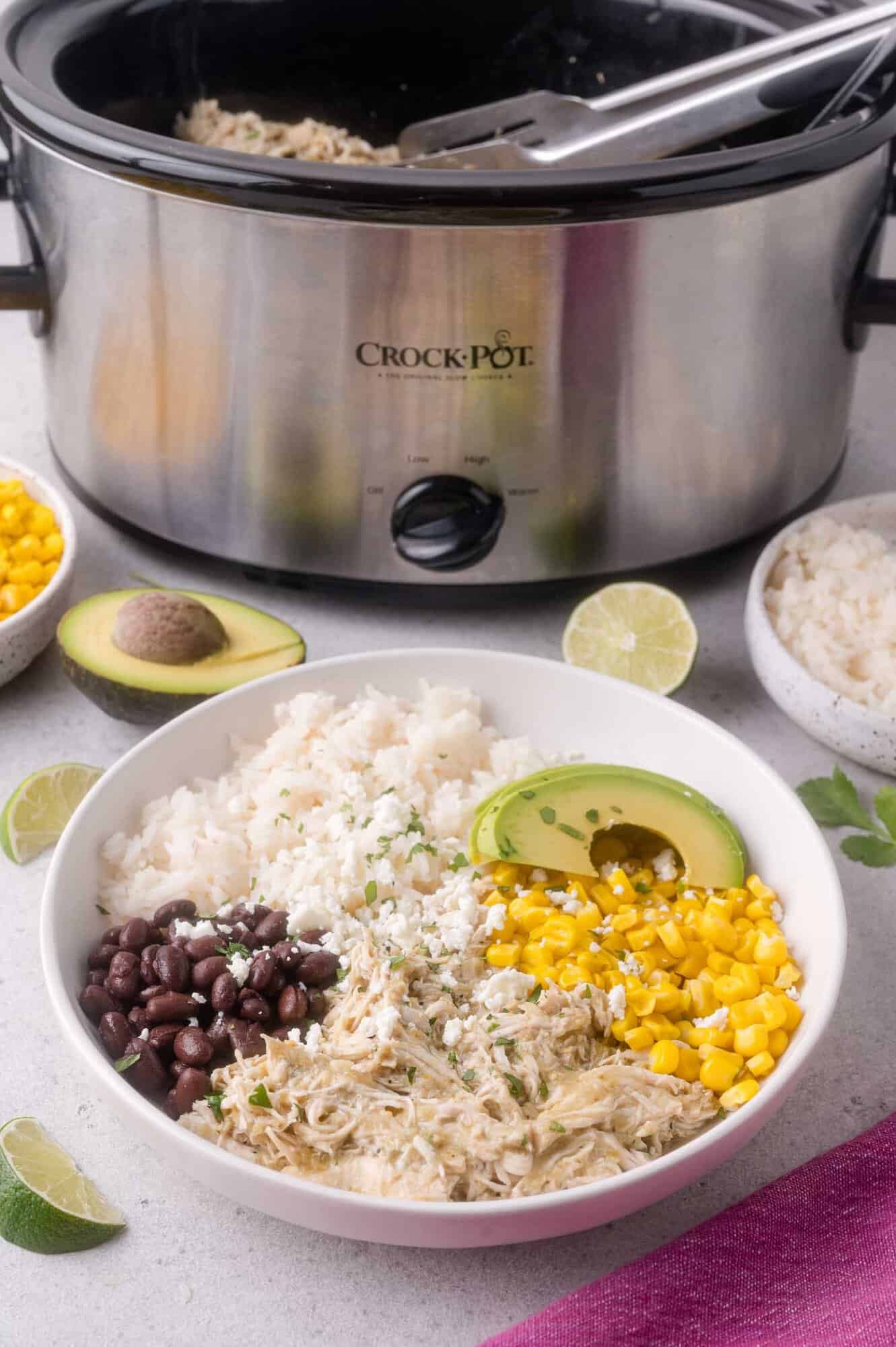 A white bowl filled with shredded chicken, white rice, black beans, corn, avocado, and cheese sits in front of a Crock-Pot slow cooker. Lime slices, rice, and corn are nearby on the table.