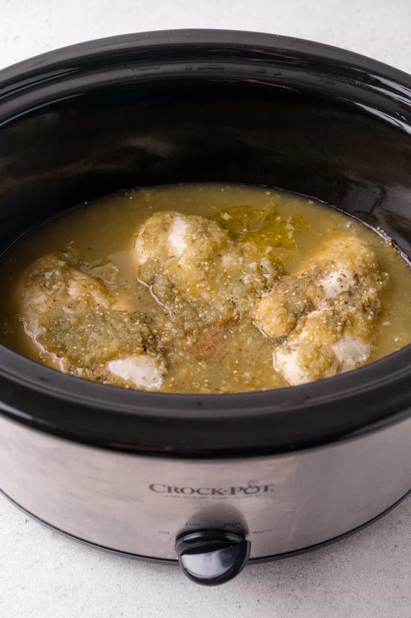 Chicken breasts cooking in green salsa inside a Crock-Pot slow cooker on a light countertop.