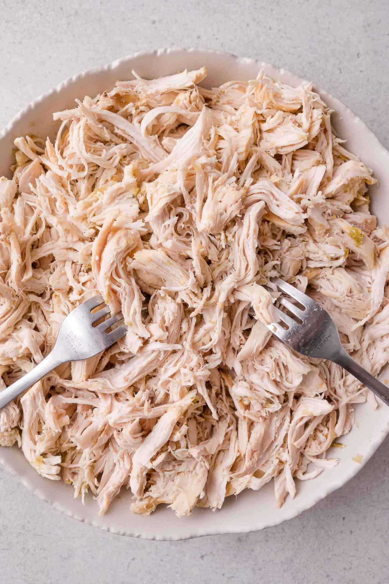 A bowl of shredded cooked chicken with two forks resting in it, on a light-colored surface.