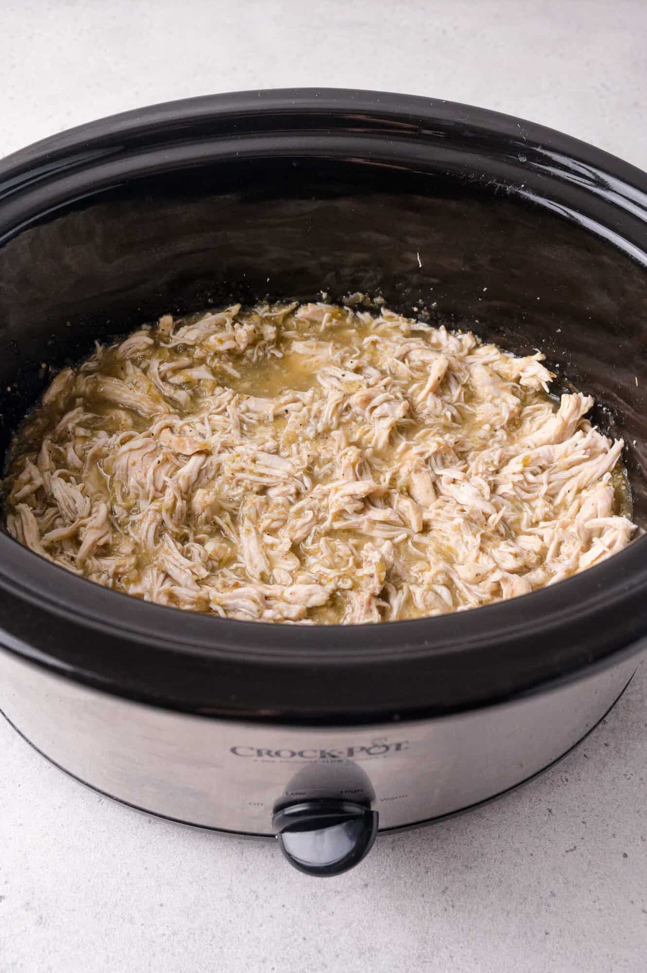Shredded chicken in salsa verde inside a black slow cooker on a light countertop.