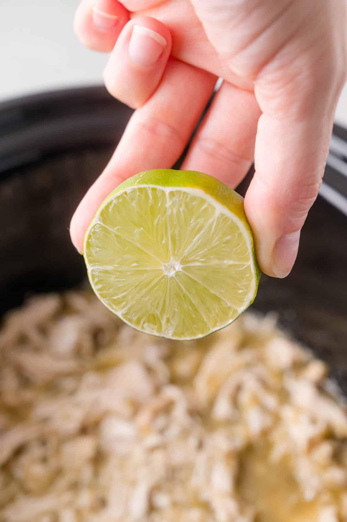 A hand holds half a lime above shredded chicken in a slow cooker, preparing to squeeze the juice onto the food.