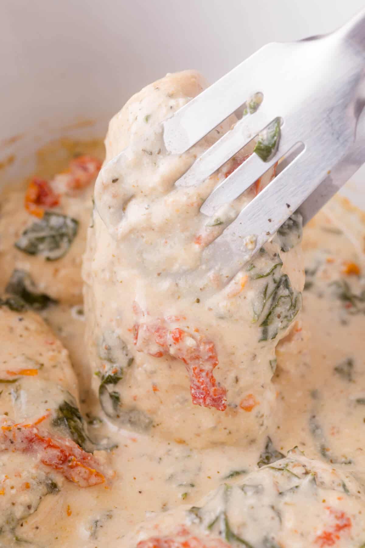 Tongs hold a piece of chicken covered in a creamy sauce with spinach and sun-dried tomatoes.