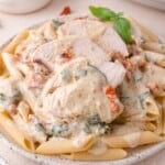 Plate of penne pasta topped with sliced chicken breast, creamy white sauce, spinach, and sun-dried tomatoes, garnished with a basil sprig.