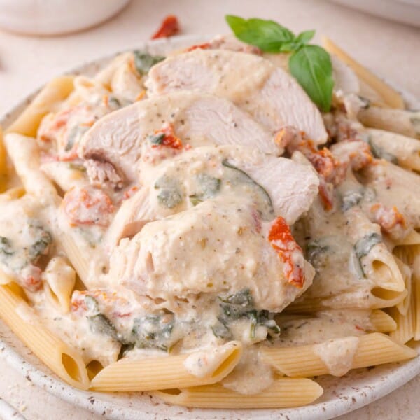 Plate of penne pasta topped with sliced chicken breast, creamy white sauce, spinach, and sun-dried tomatoes, garnished with a basil sprig.