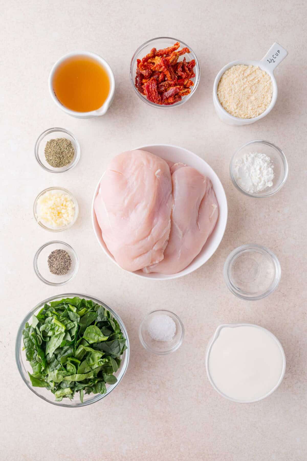 Top-down view of chicken breasts, fresh spinach, cream, sun-dried tomatoes, parmesan cheese, garlic, salt, pepper, Italian seasoning, and cornstarch in individual bowls on a countertop.