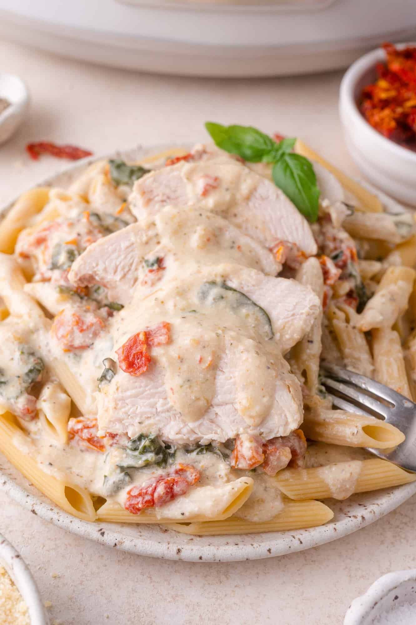 A plate of penne pasta topped with creamy sauce, sliced chicken breast, sun-dried tomatoes, and spinach, garnished with a basil leaf.