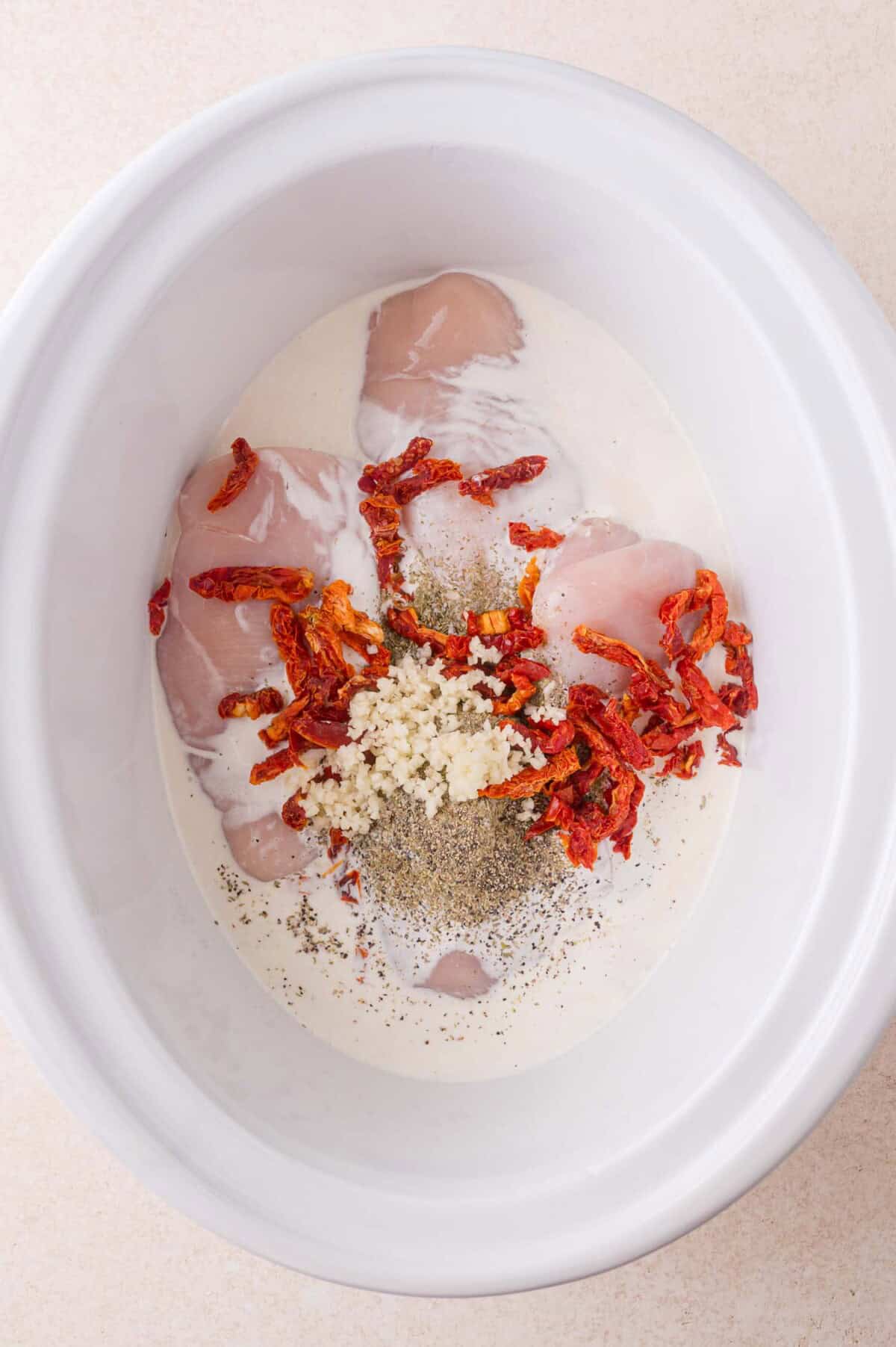 Raw chicken breasts, cream, sun-dried tomatoes, minced garlic, and black pepper are placed in a white slow cooker before mixing.