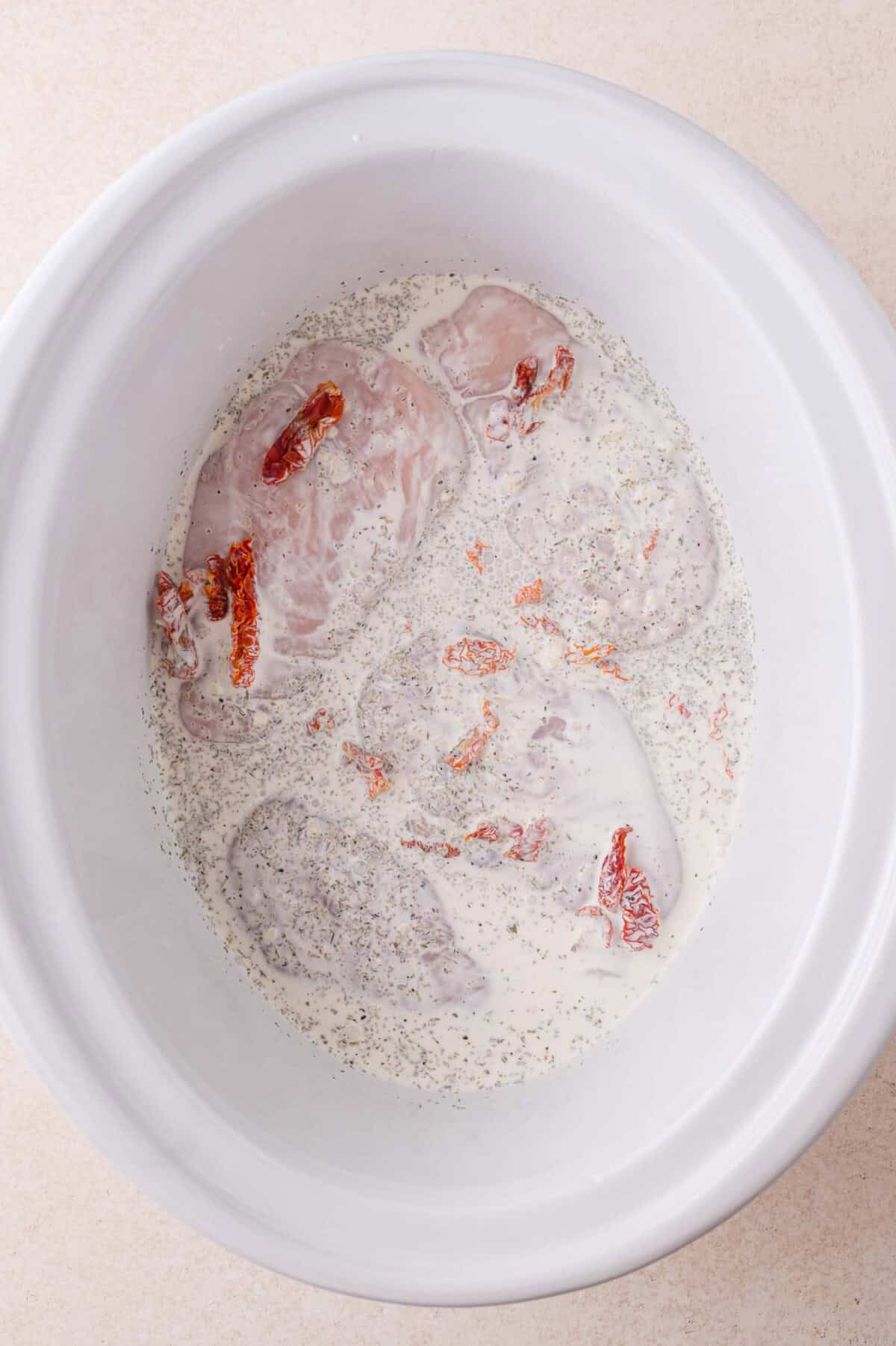 Raw chicken breasts in a white slow cooker with cream, black pepper, and sun-dried tomato pieces.